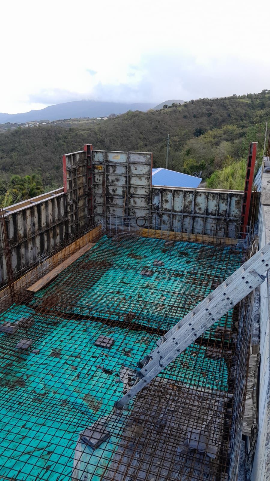 Chantier de construction avec des murs de coffrage métallique et un treillis d'armature étalé sur le sol à flanc de colline offrant une vue panoramique.