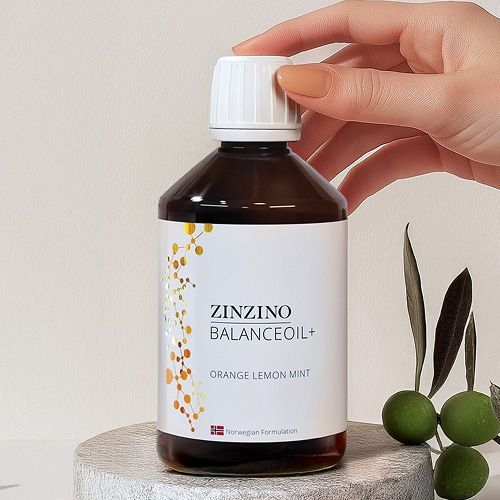 Una mano coloca la tapa en la botella ámbar de aceite Zinzin Balance, con aceitunas y hojas cerca.
