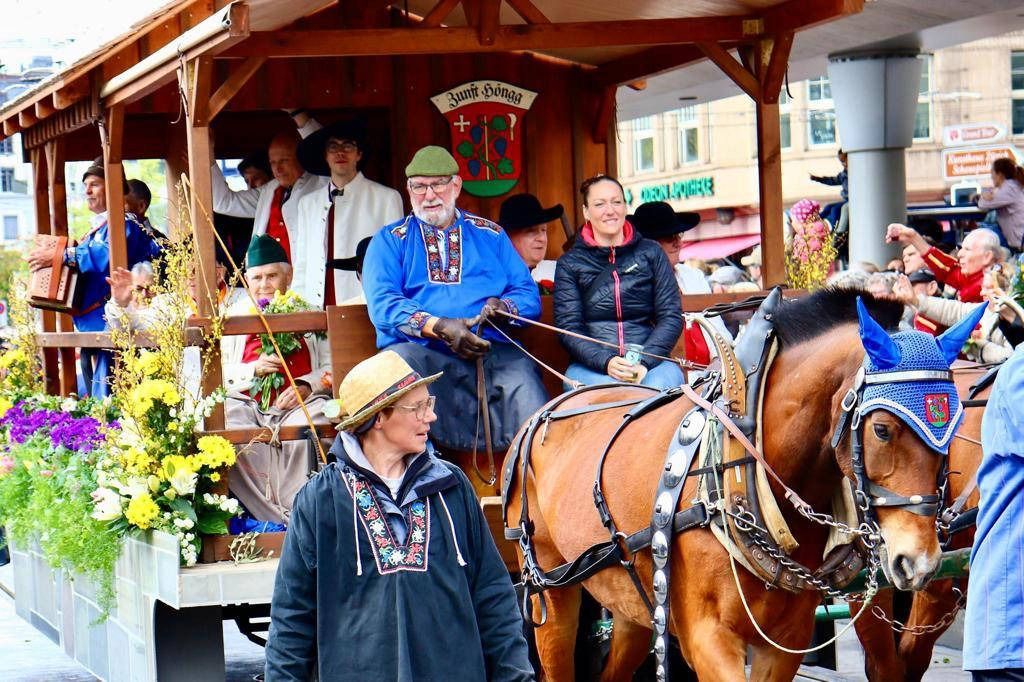 Eine von Pferden gezogene Kutsche in einer Parade mit Menschen in traditioneller Kleidung, bunten Blumen und Zuschauern im Hintergrund.