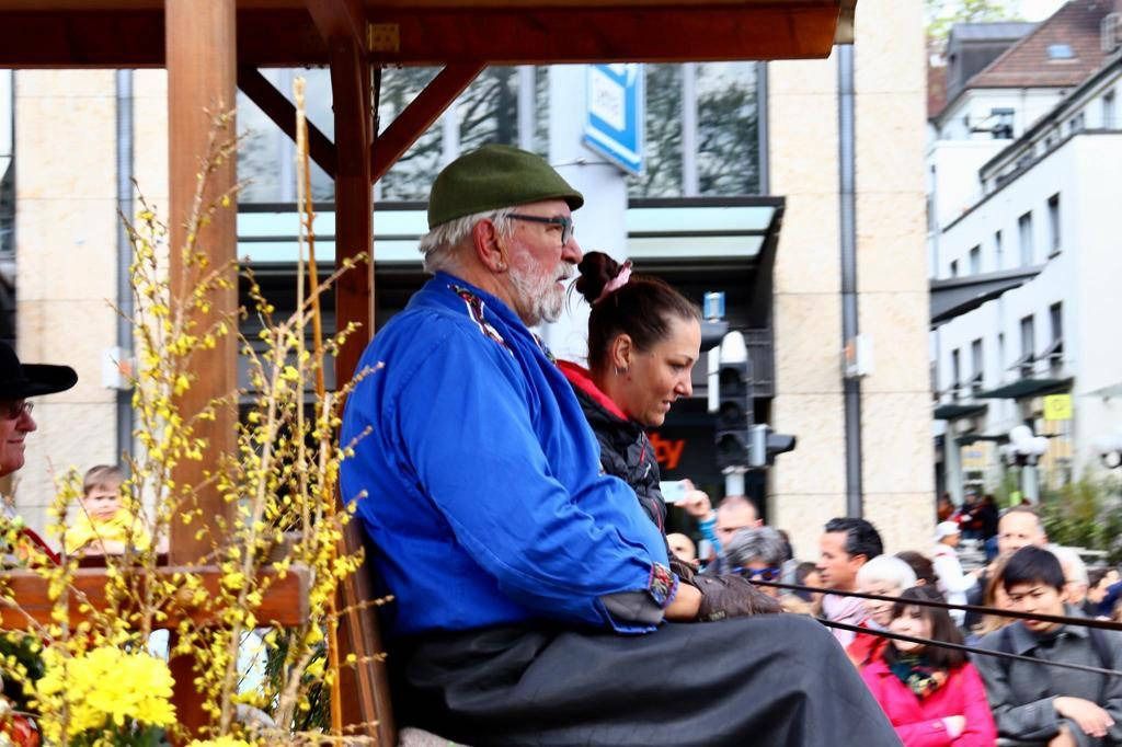 Ein Mann in blauer Jacke und grünem Hut sitzt auf einem Festwagen und beobachtet eine Parade. Im Hintergrund sind weitere Personen zu sehen.
