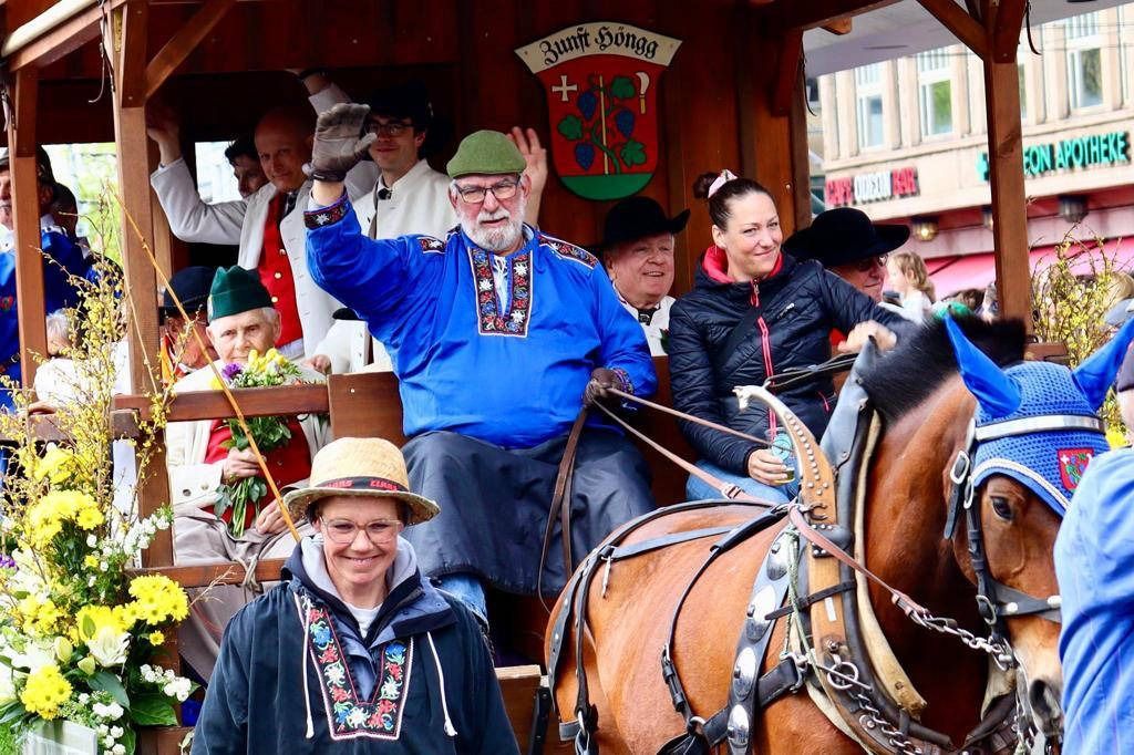 Menschen auf einem Pferdewagen winken, feiern und lächeln. Im Hintergrund ist das Stadtwappen auf dem Wagen zu sehen.
