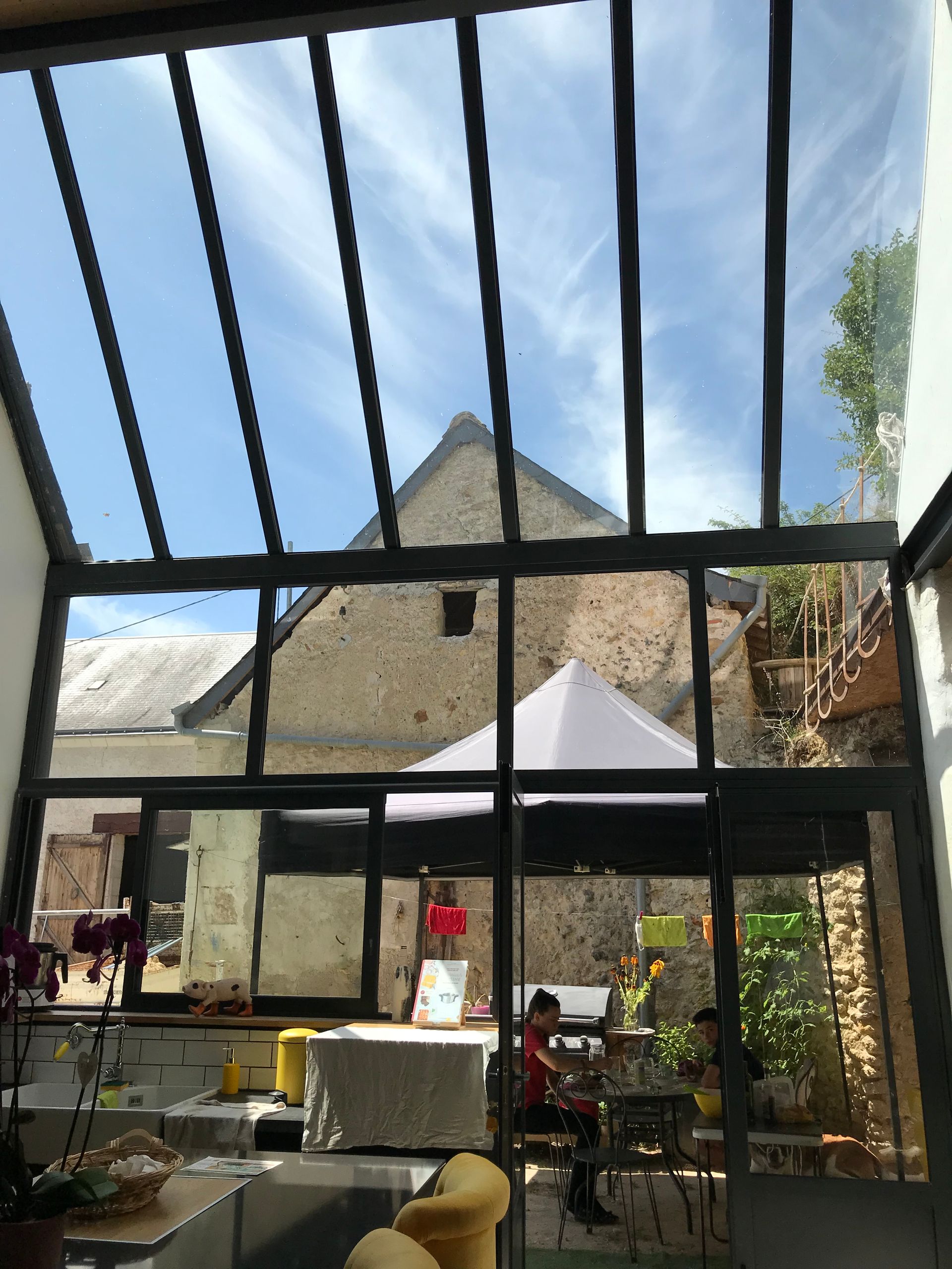 Véranda noire avec fenêtres et porte vitrées, attenante à une maison beige, terrasse en pierre et pelouse verte.