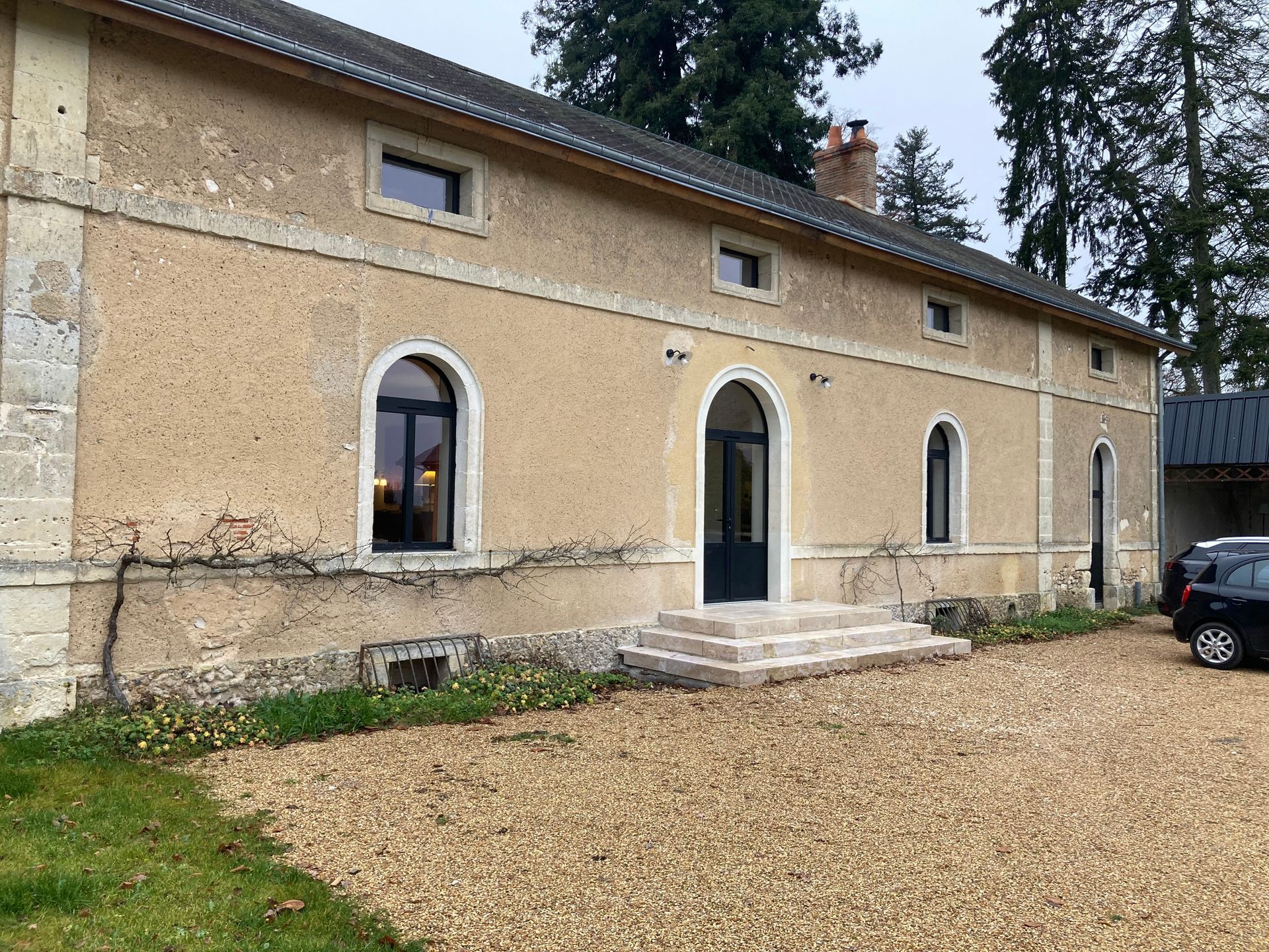 Bâtiment ancien avec fenêtres à encadrement foncé, cour gravillonnée, marches en pierre et porte en aluminium gris foncé.