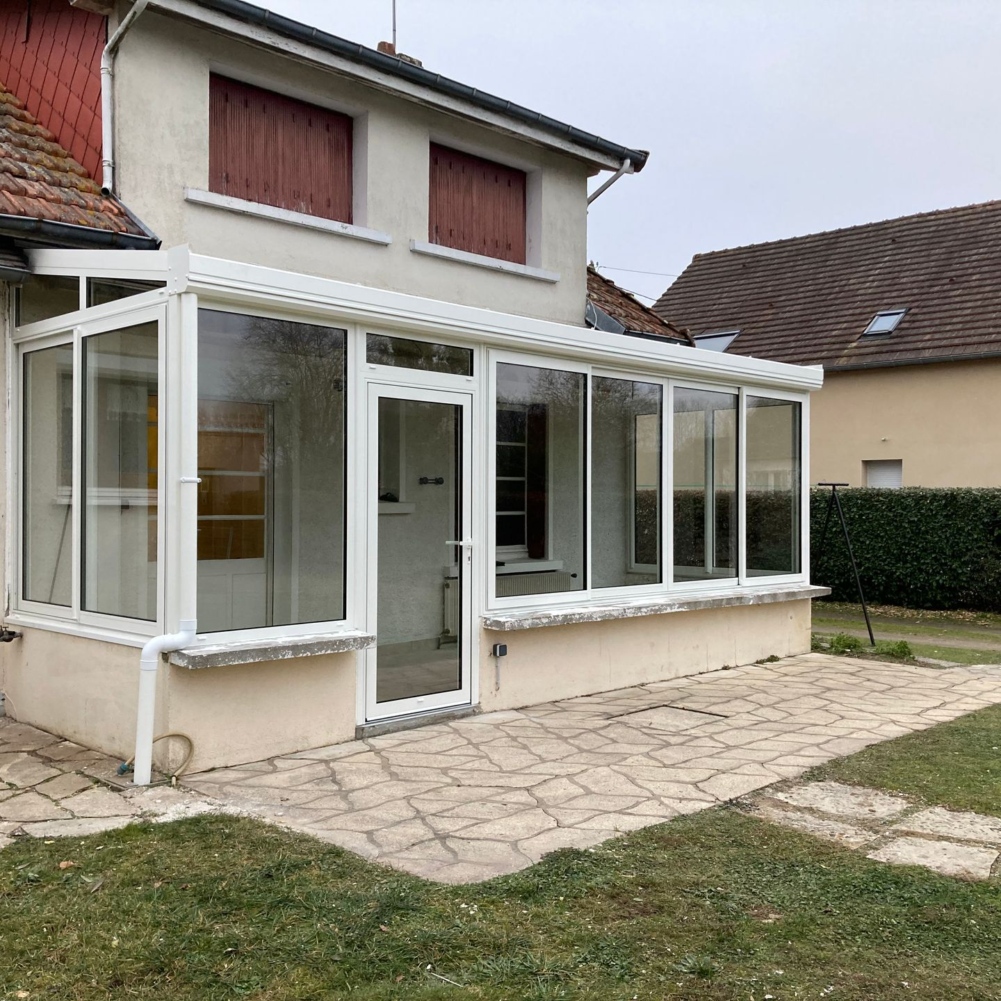 Véranda blanche avec fenêtres et porte vitrées, attenante à une maison beige, terrasse en pierre et pelouse verte.