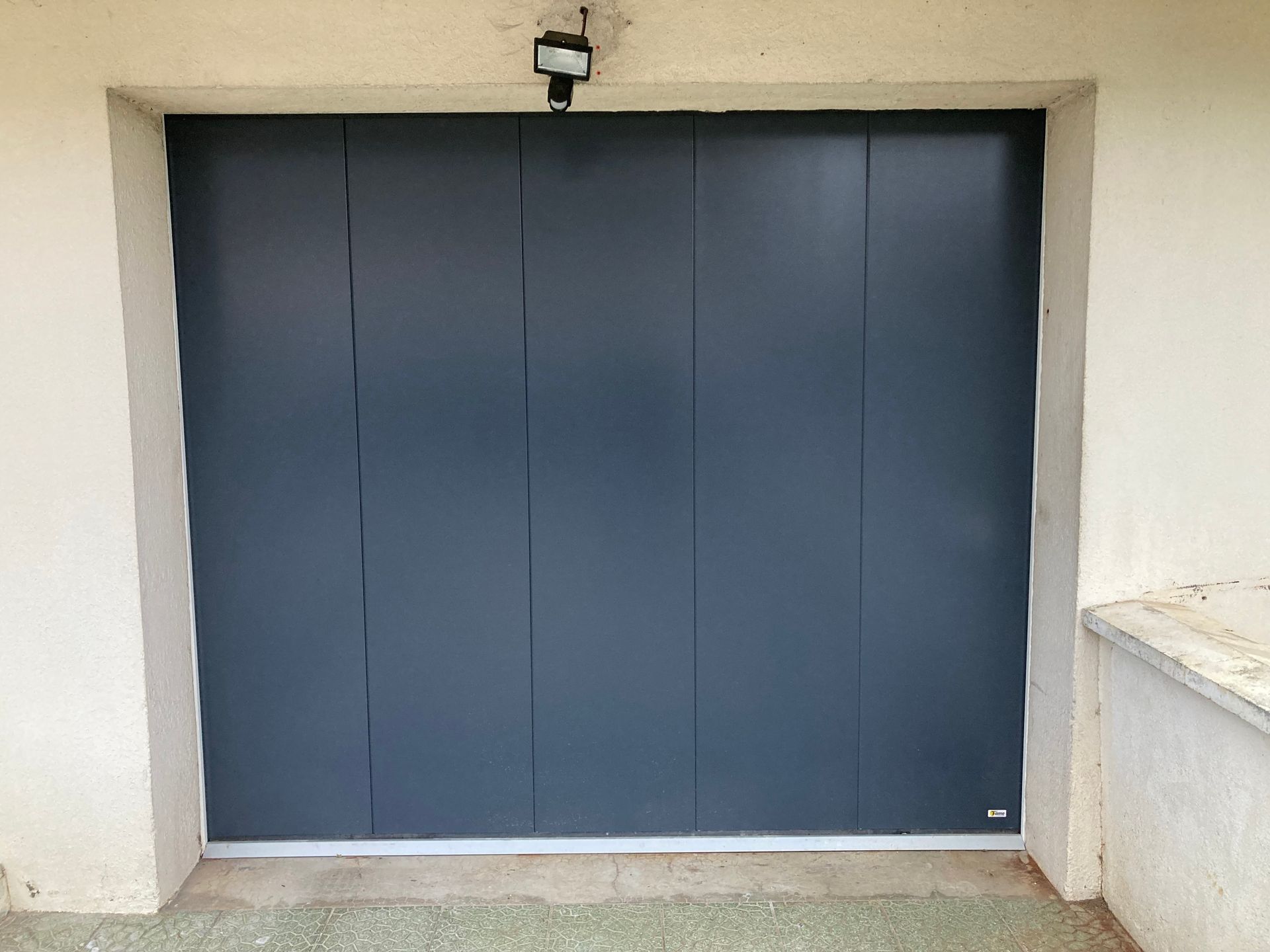 Porte de garage gris foncé avec cadre blanc cassé. Un détecteur de mouvement est installé au-dessus de la porte.