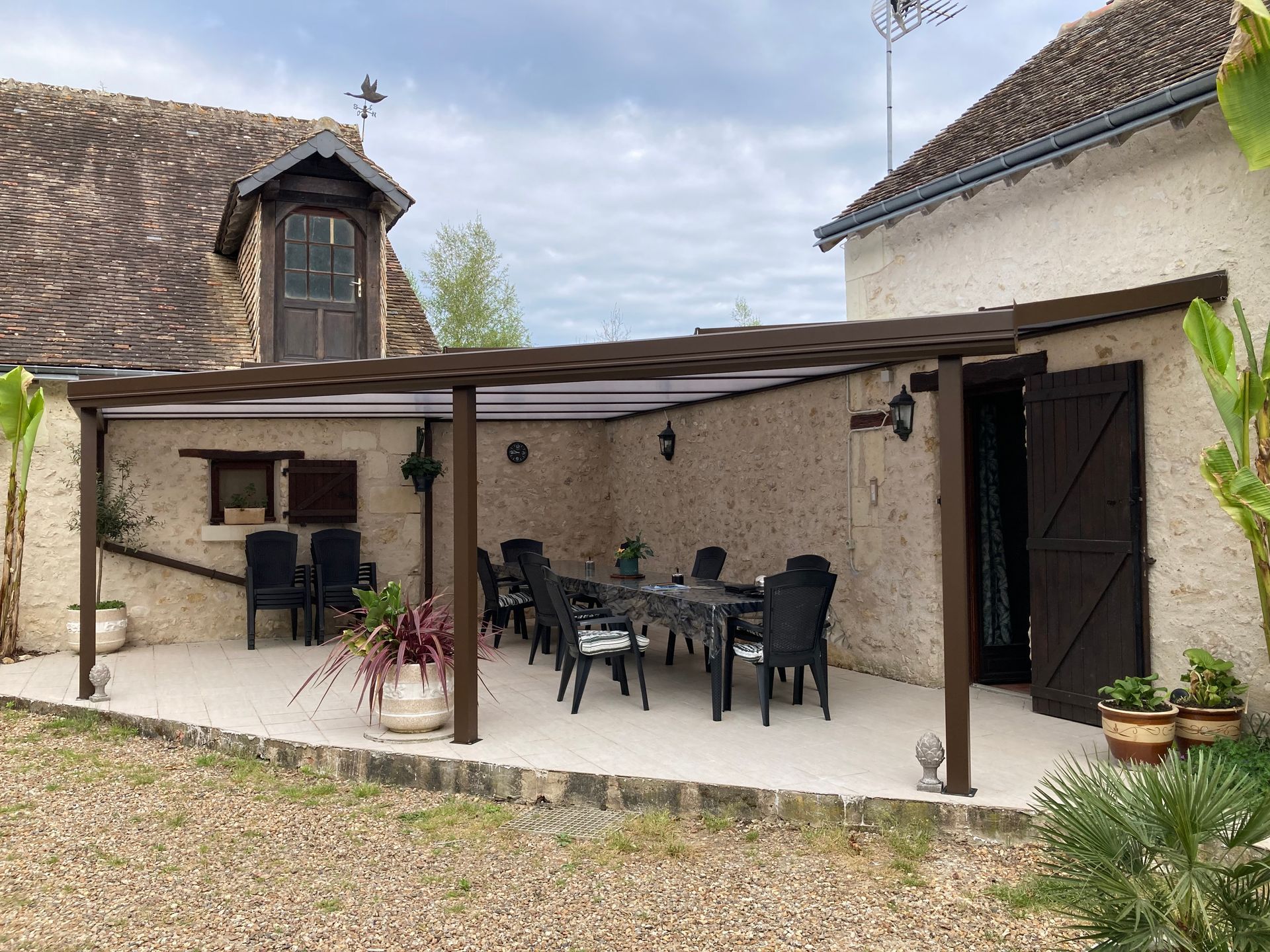 Terrasse avec table à manger sous une pergola marron, à côté d'un bâtiment en pierre aux volets marron.