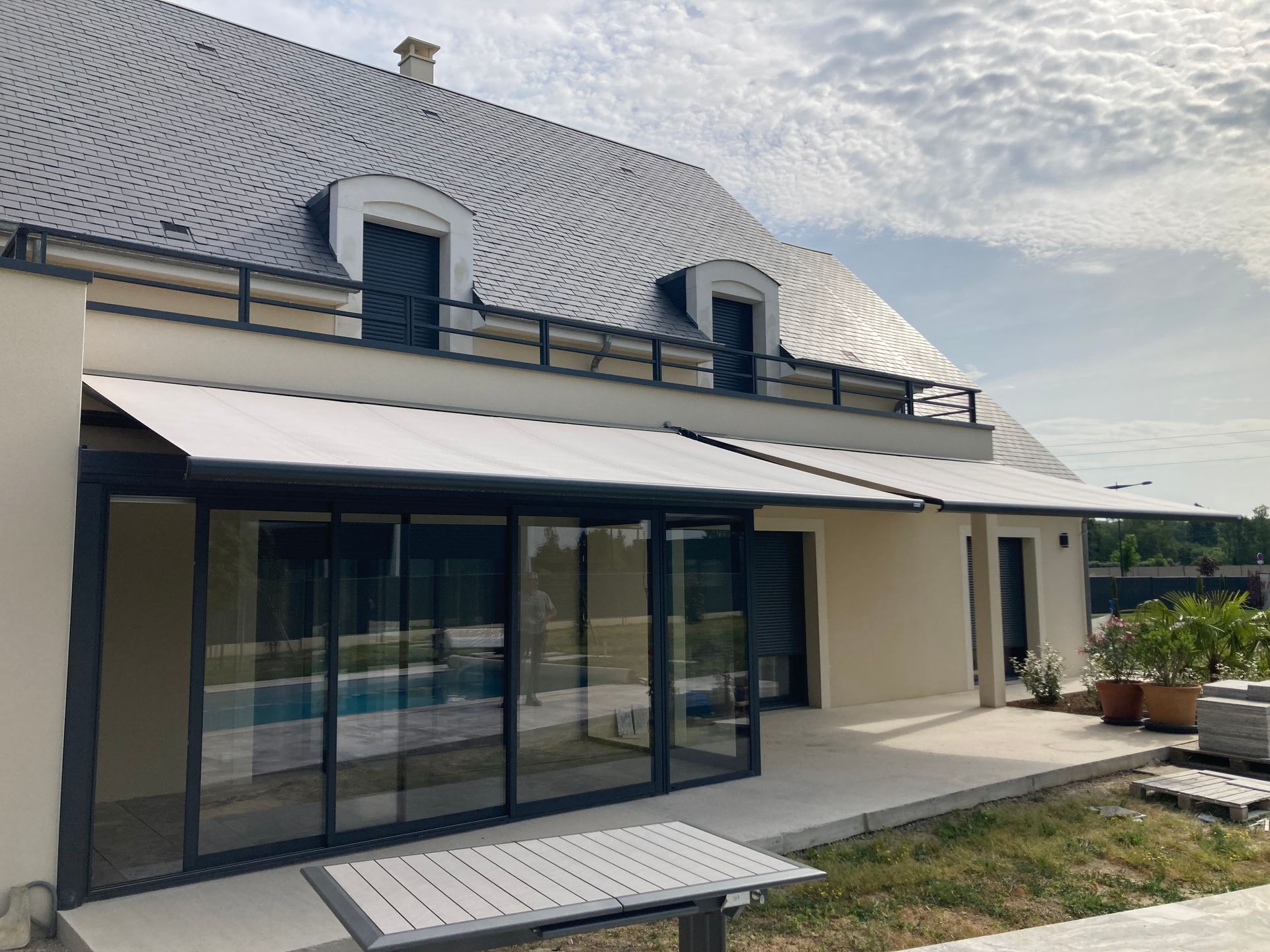 Maison moderne avec store banne de couleur claire au-dessus d'une terrasse bordée par une piscine.