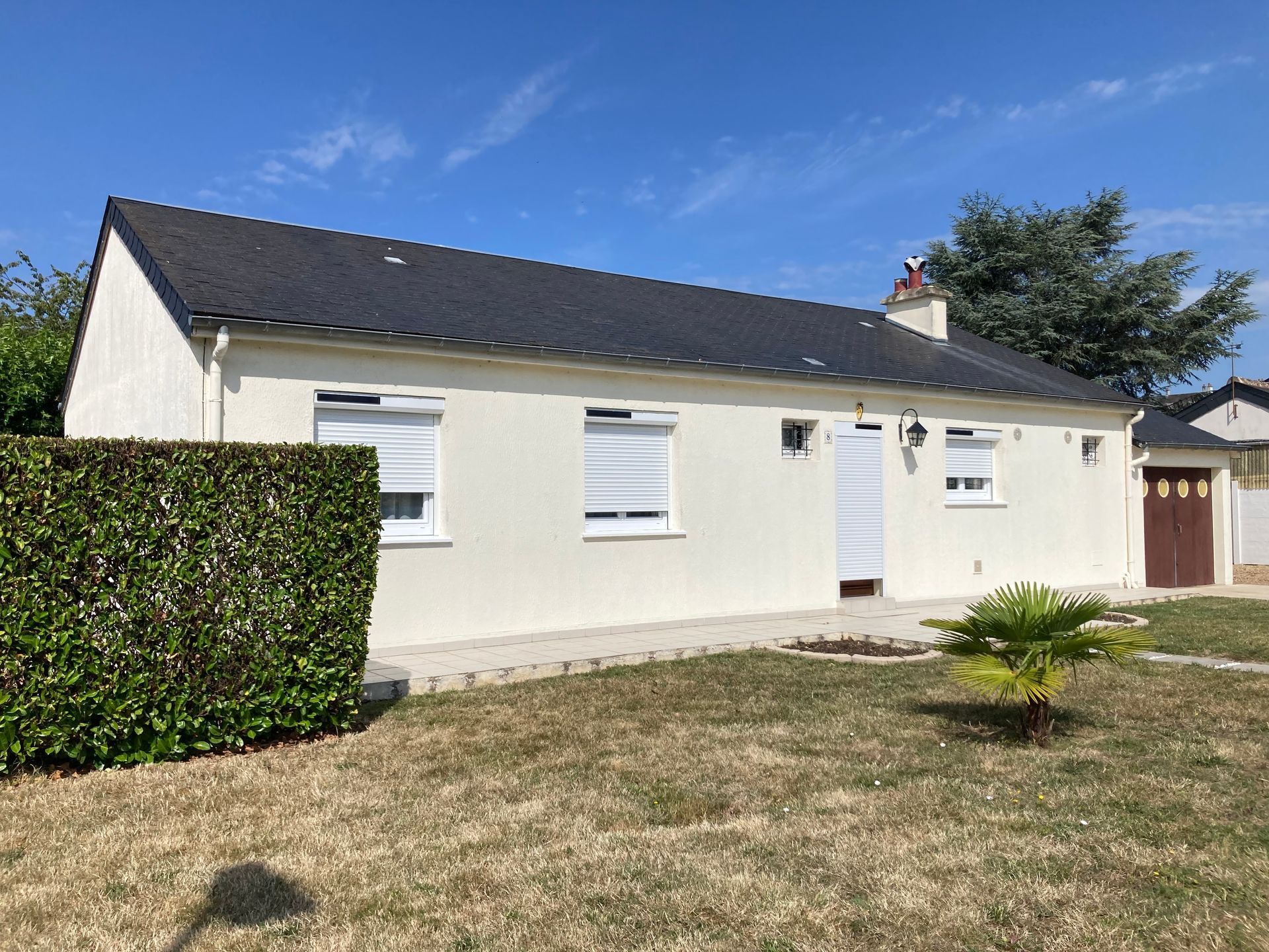 Maison beige de plain-pied avec toit foncé et volets blancs, située sur une pelouse sous un ciel bleu.