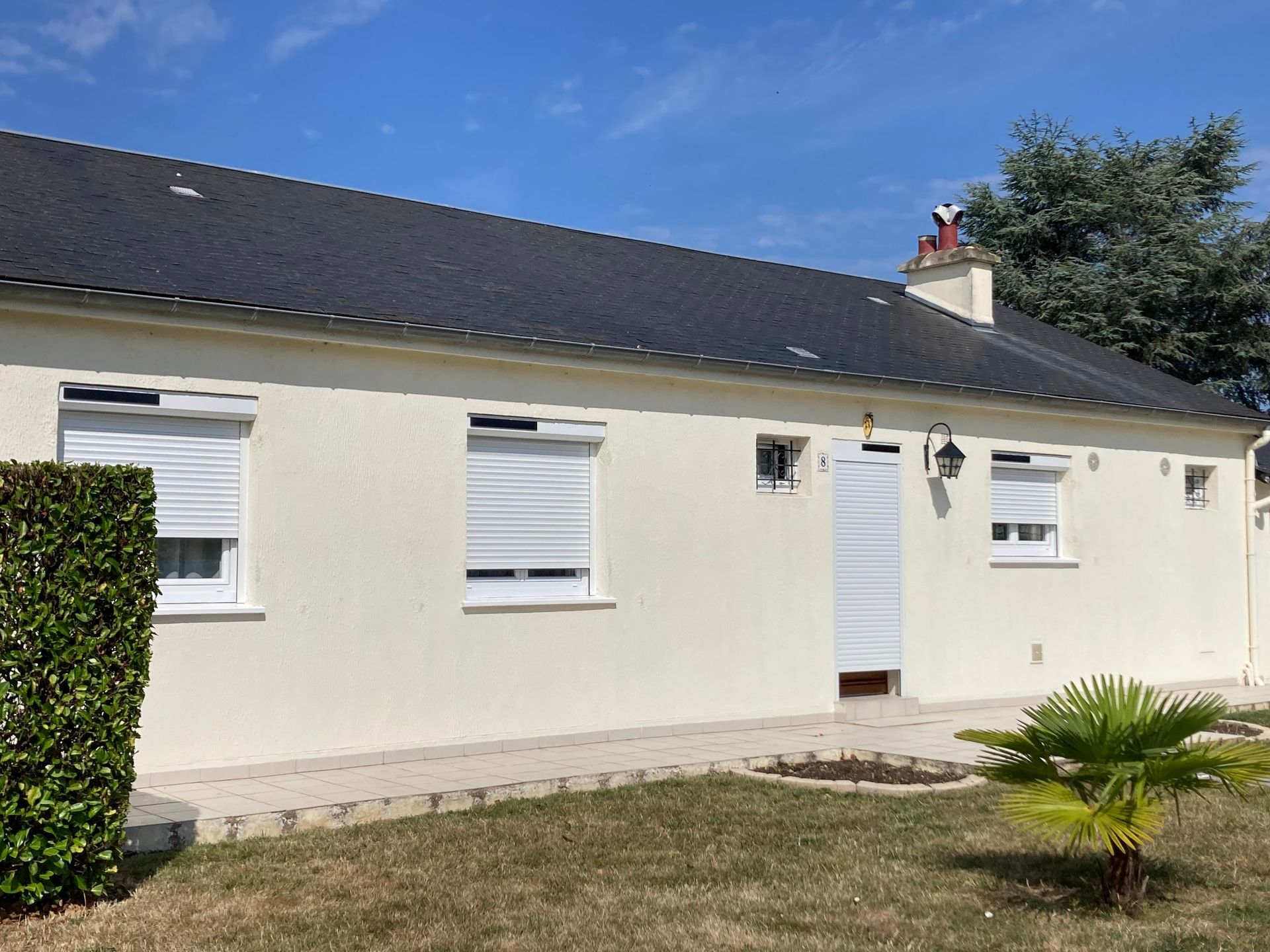 Maison beige avec toit noir, volets blancs et petit jardin sous un ciel bleu.