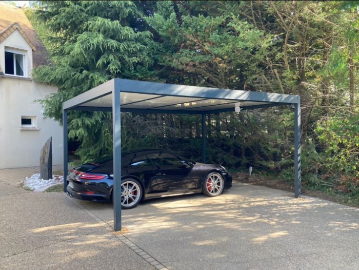 Une voiture de sport noire est garée sous un carport gris dans une allée pavée, près d'une maison et d'arbres.