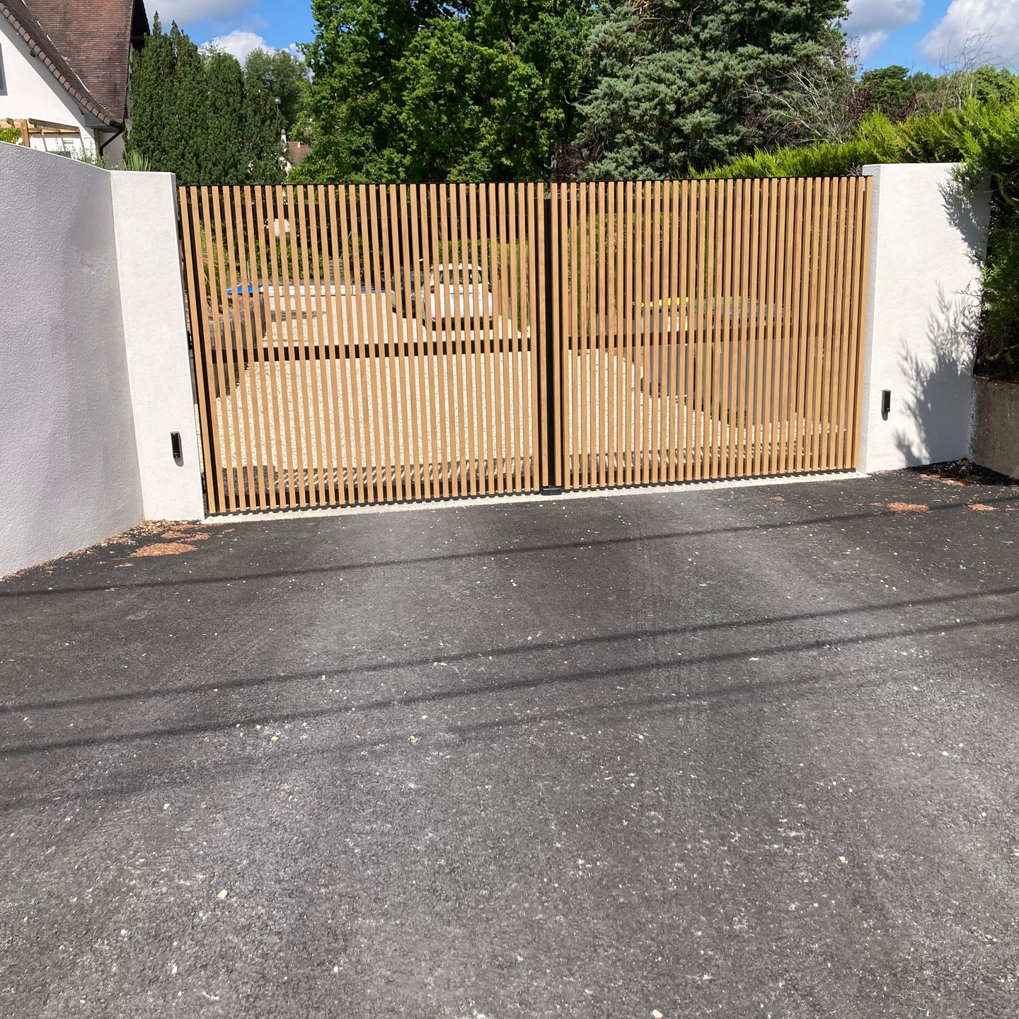 Portail en bois à lattes donnant sur une allée, flanqué de piliers blancs et d'une surface asphaltée.