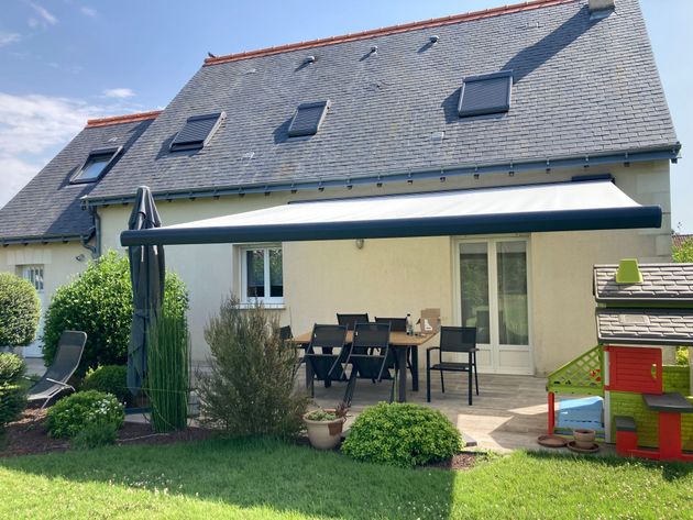Une terrasse avec un store banne ouvert, une table et des chaises devant une maison au toit d'ardoise et une petite cabane de jeux.