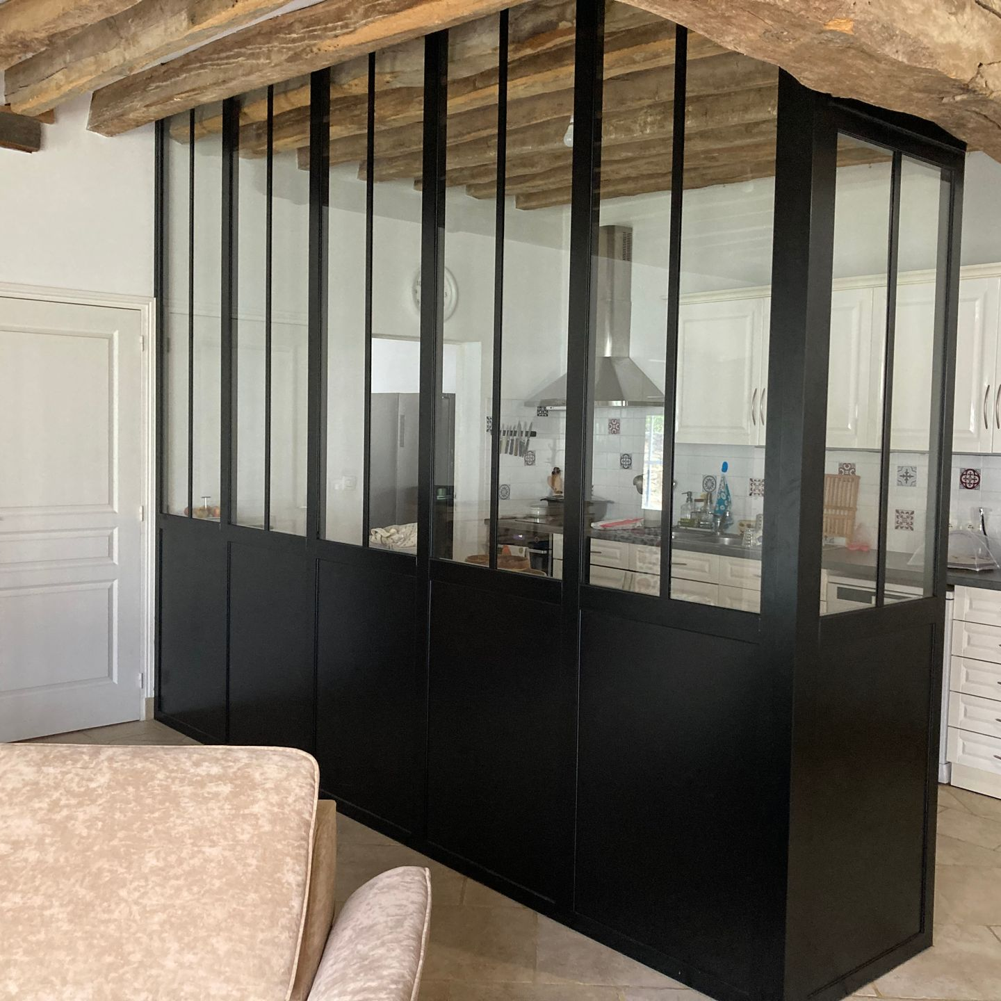Une verrière en aluminium noir sépare un espace de vie d'une cuisine, avec des poutres en bois apparentes au-dessus.