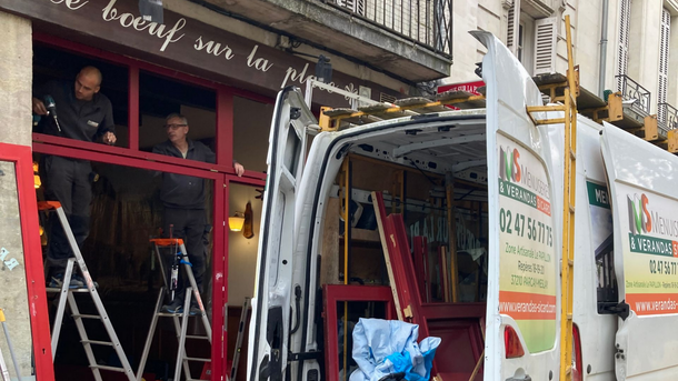 Des ouvriers installent un cadre de porte rouge sur un bâtiment. Une camionnette est garée devant, on aperçoit les outils.
