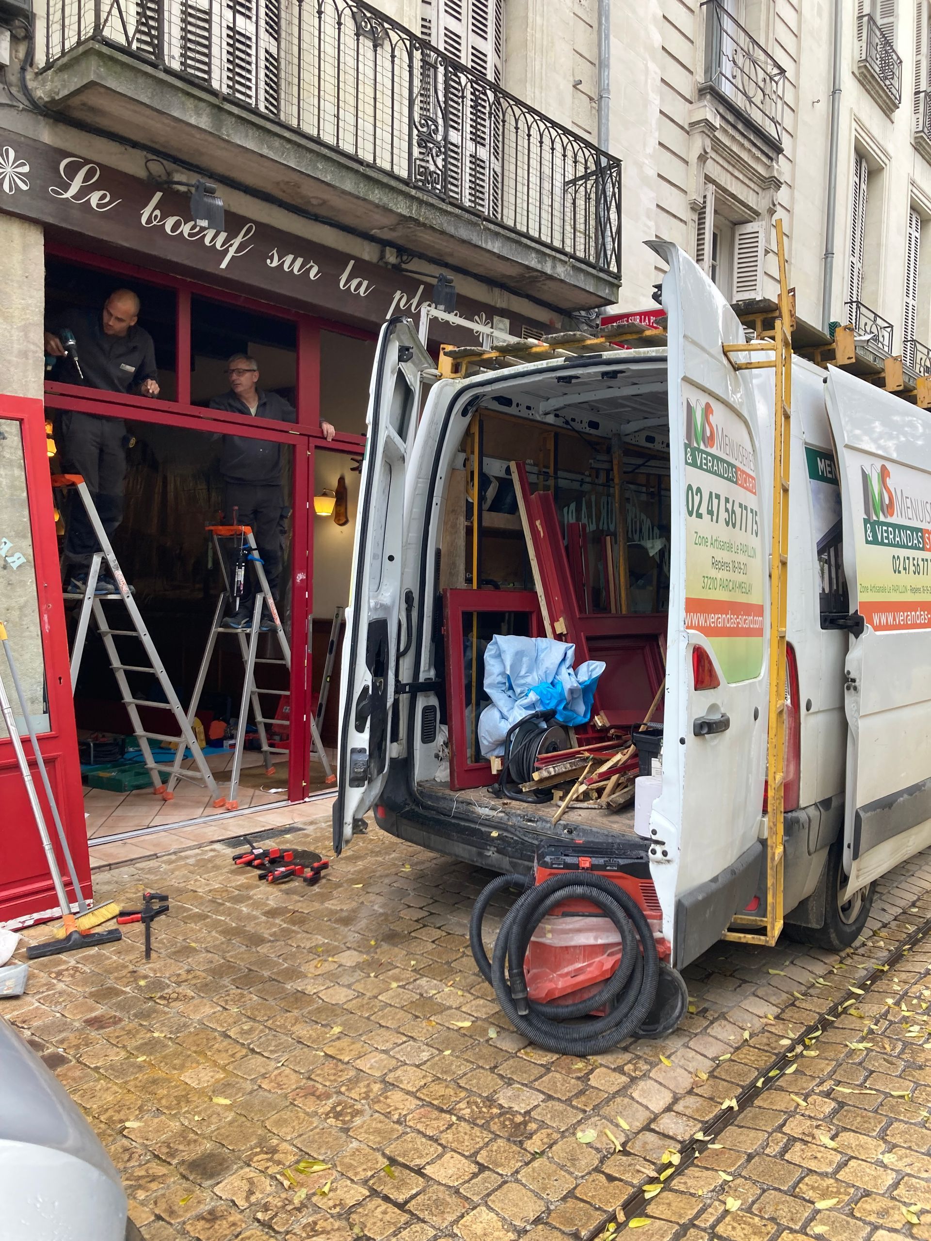 Des ouvriers installent un cadre de porte rouge sur un bâtiment. Une camionnette est garée devant, on aperçoit les outils.