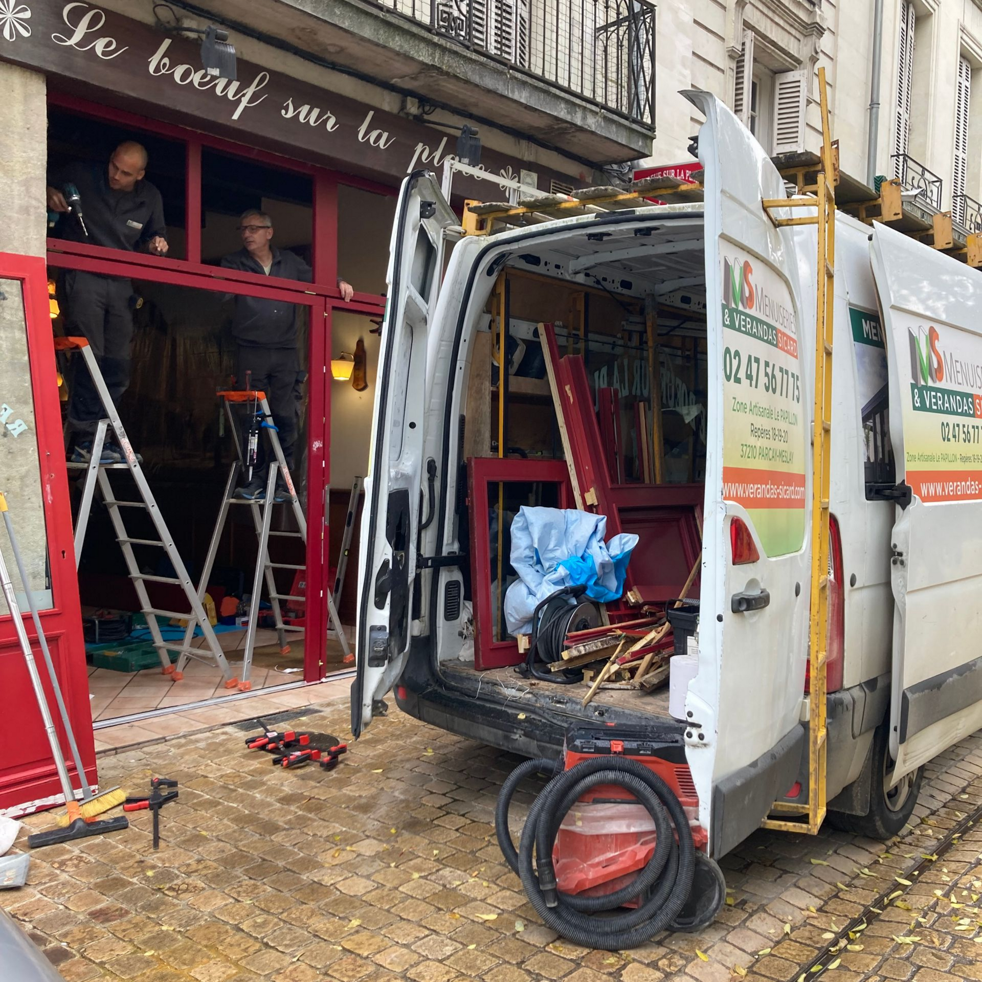 Des ouvriers installent un cadre de porte rouge sur un bâtiment. Une camionnette est garée devant, on aperçoit les outils.