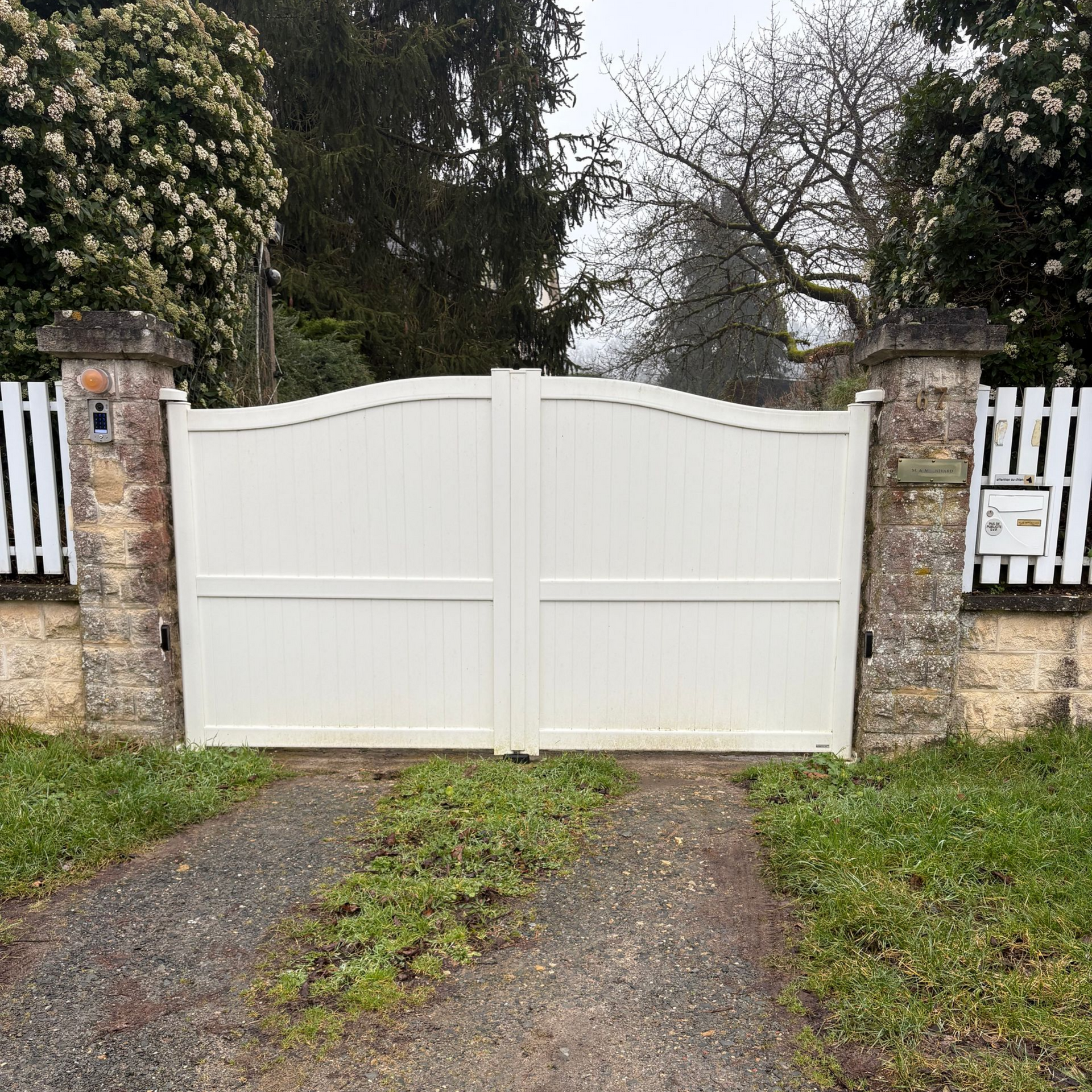 Un double portail blanc entre des piliers de pierre, fermé sur une allée de gravier, avec des arbres en arrière-plan.