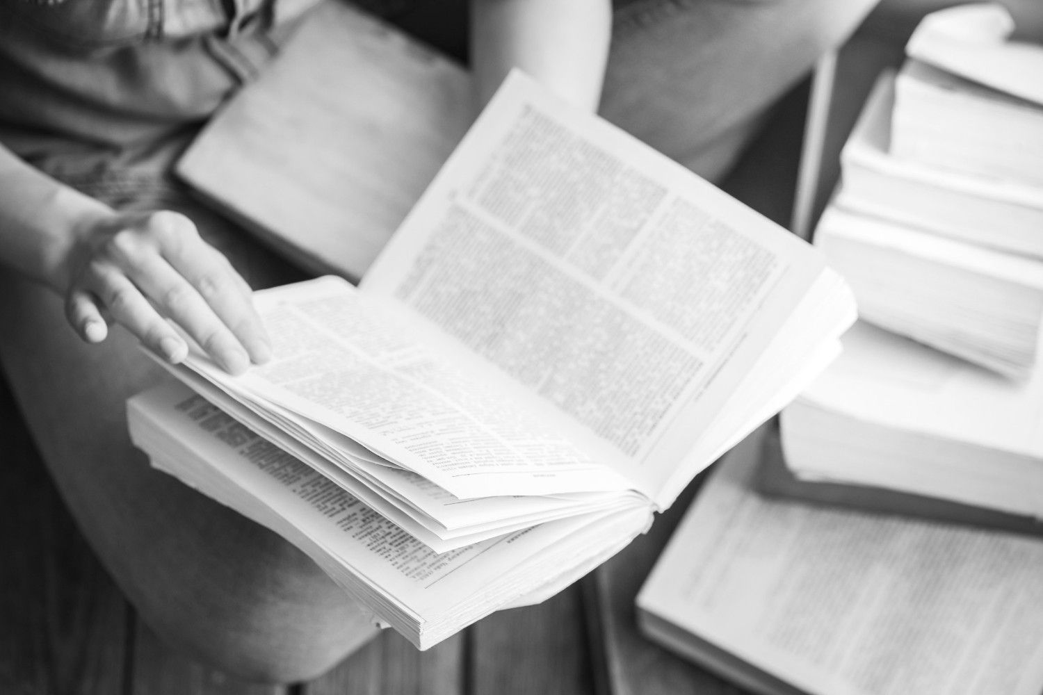 Persona hojeando un libro abierto, rodeada de otros libros sobre una superficie de madera.