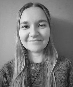 Una mujer con cabello largo y rubio sonríe, vistiendo un suéter gris.