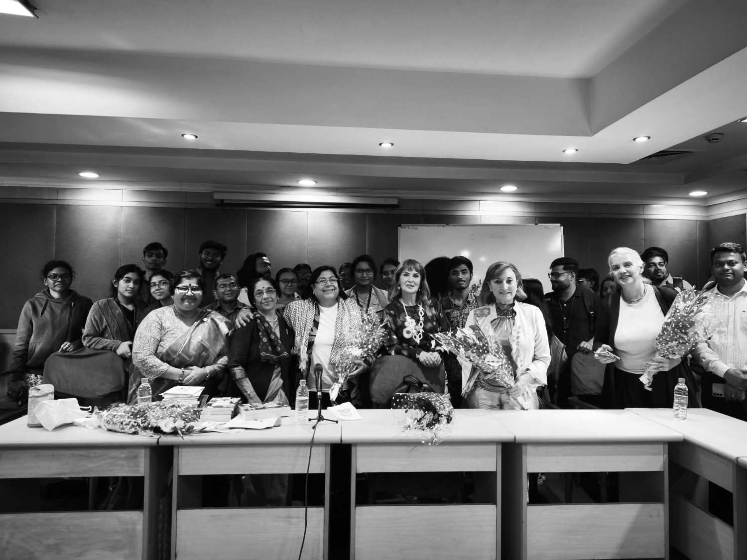 Dos personas están de pie junto a una pancarta y un logotipo. El hombre lleva una camisa clara y pantalones marrones. La mujer lleva una camisa rosa y pantalones negros.