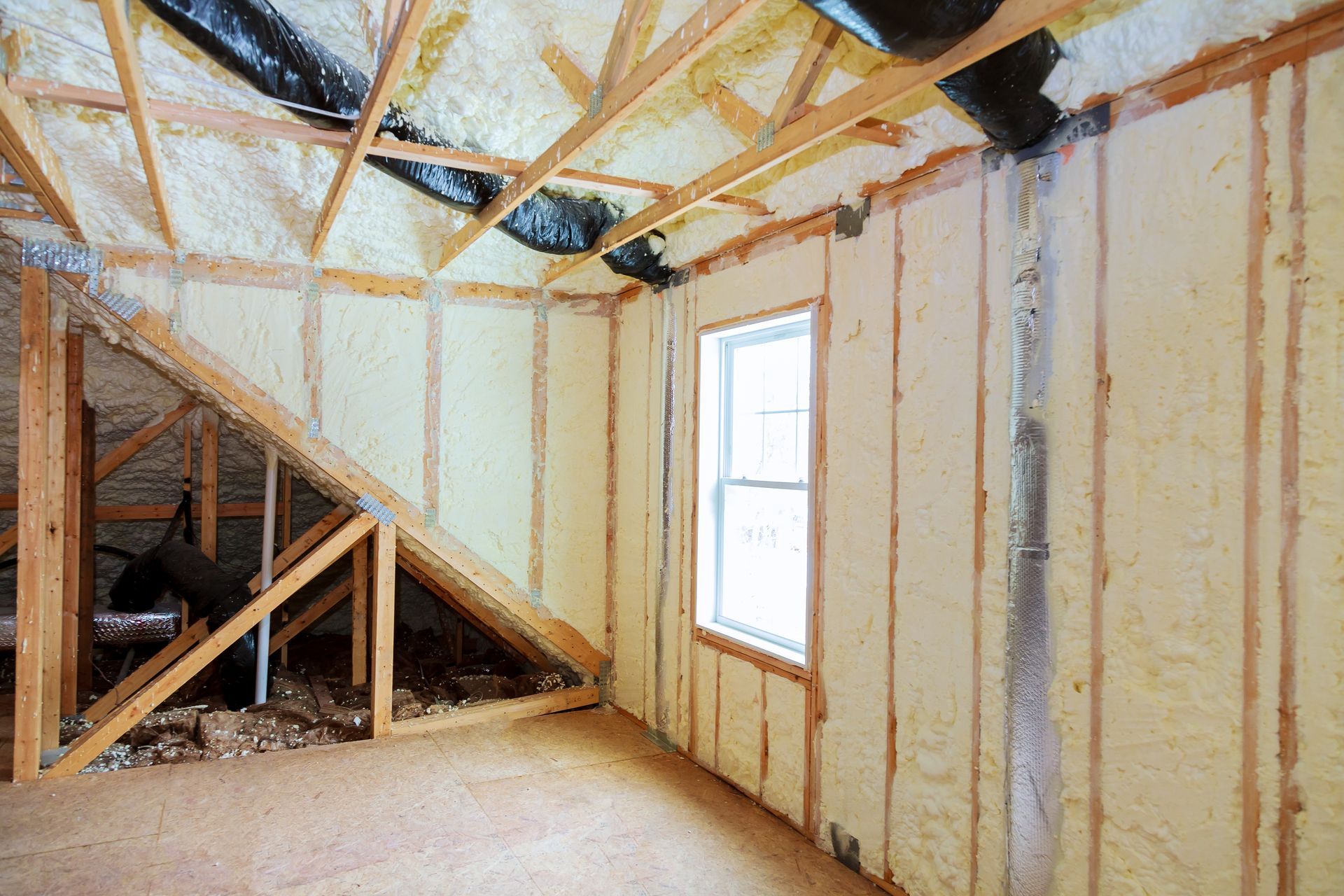 Intérieur de grenier non aménagé avec charpente apparente, isolation et une petite fenêtre laissant passer la lumière.