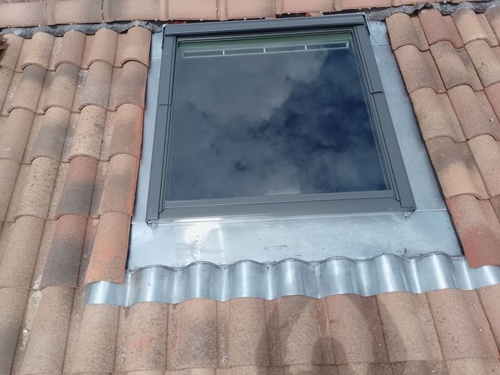 Puits de lumière sur le toit, entouré de tuiles en terre cuite rouge et de solins, reflétant les nuages ​​dans le verre.