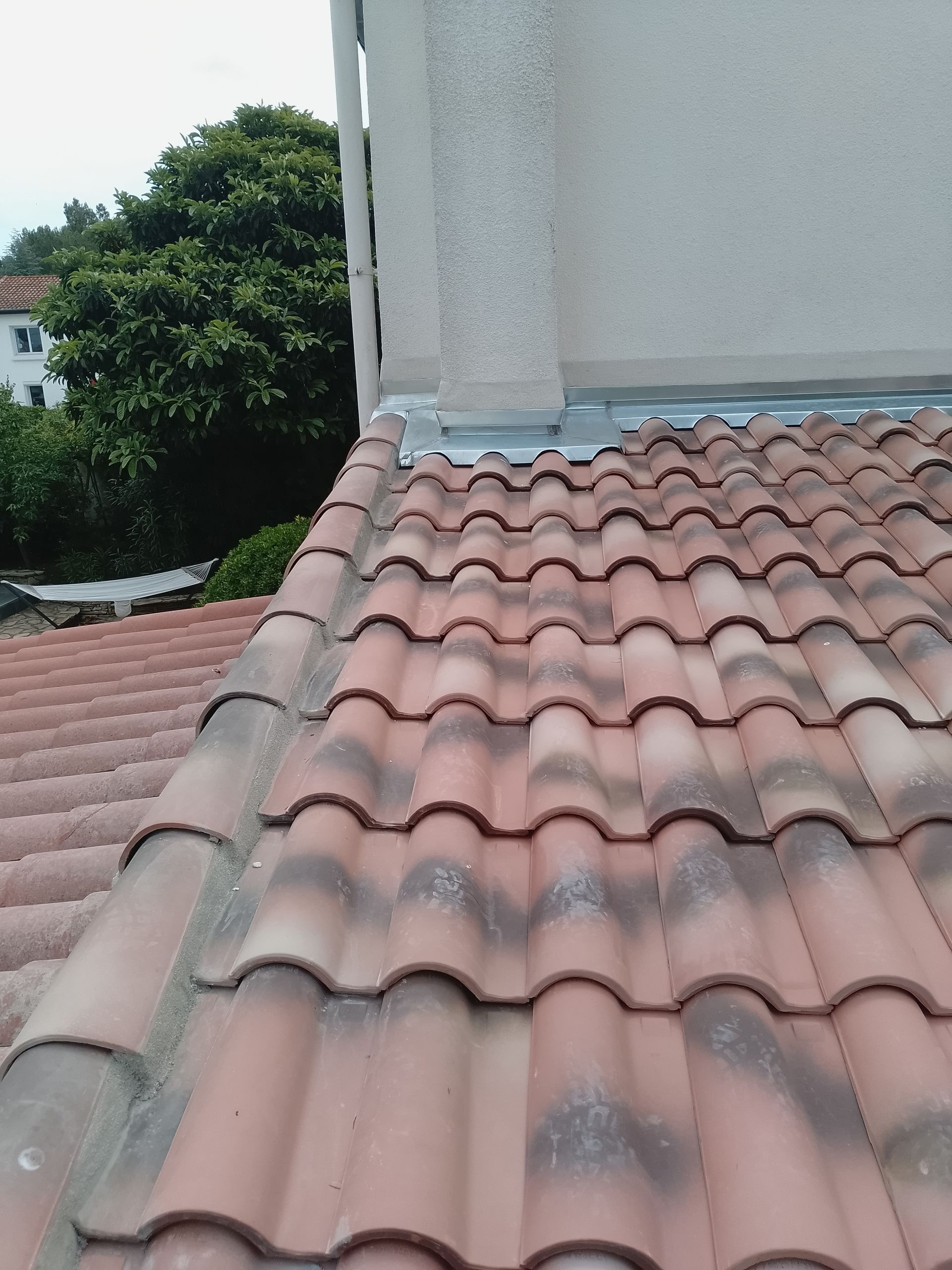 Des tuiles en terre cuite sur un toit, à côté d'un mur extérieur blanc, avec un arbre vert en arrière-plan.