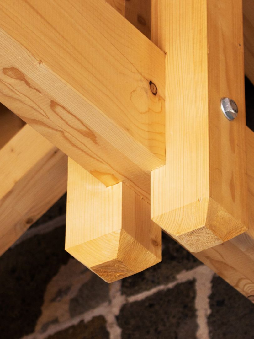 Gros plan sur une structure en bois avec des poutres inclinées et un boulon métallique sur un sol à motifs sombres.