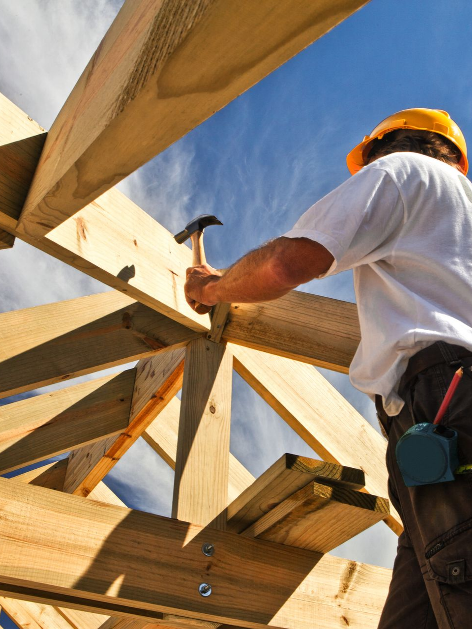 Un ouvrier du bâtiment, casque de chantier sur la tête, charpente en bois sur fond de ciel bleu.