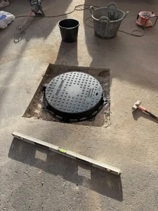 Escena de obras viales con una tapa de alcantarilla redonda colocada en asfalto recién extendido y herramientas cerca