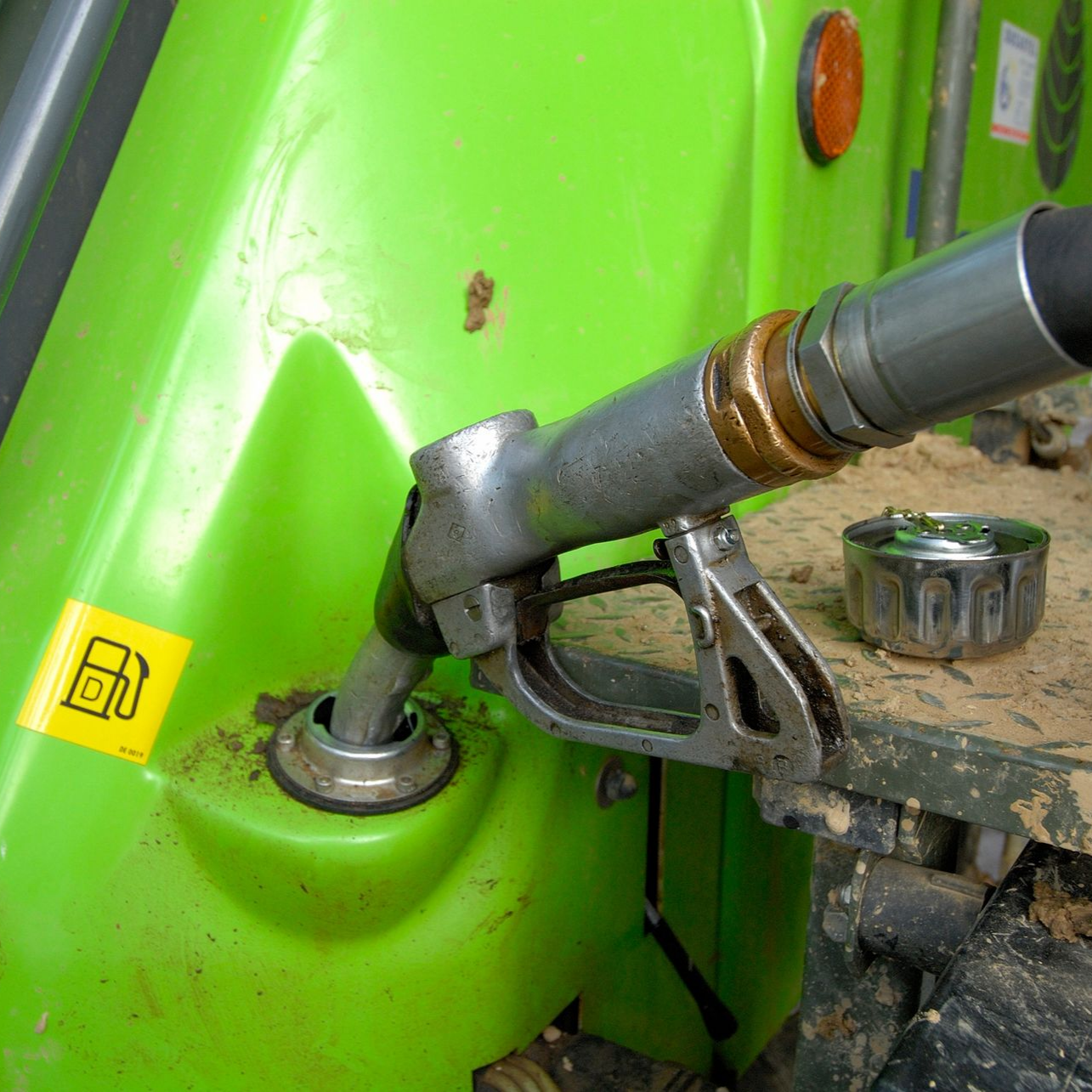 Buse de carburant remplissant le réservoir d'un véhicule vert ; bouchon de réservoir à proximité, avec un autocollant représentant une icône de pompe à carburant.