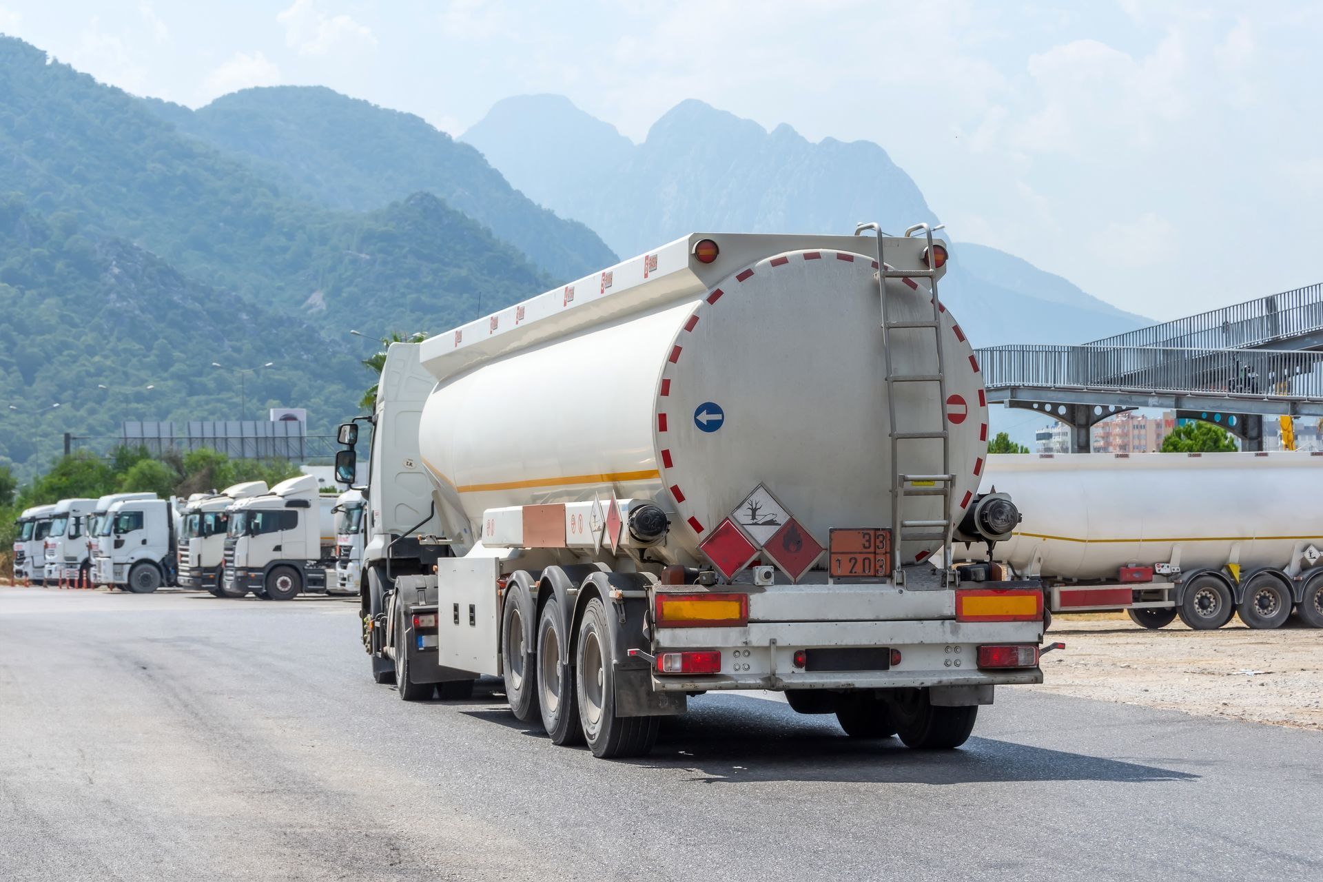 Camion-citerne blanc sur la route, avec des montagnes en arrière-plan et d'autres camions garés à proximité.