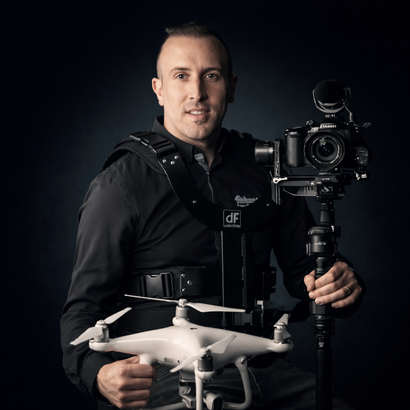 Hombre sosteniendo un dron blanco junto a una cámara montada en un estabilizador contra un fondo oscuro.