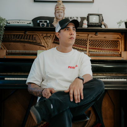 Persona con camiseta blanca y gorra negra sentada en un banco de piano frente a un piano vertical y una estatua de Buda.