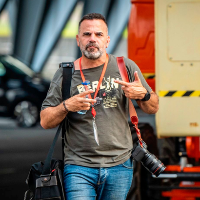 Fotógrafo con dos cámaras, vestido con camisa gris y vaqueros, de pie en un entorno industrial.