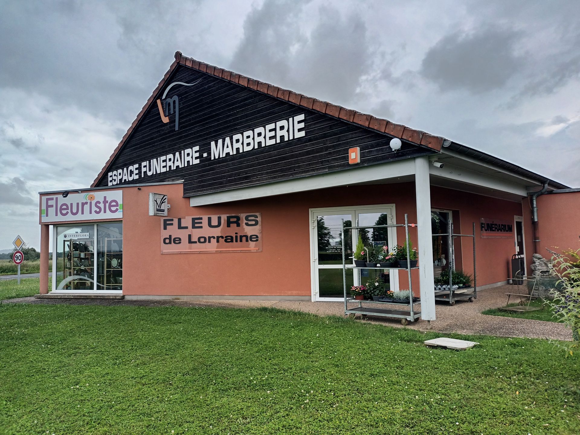 Un magasin de fleurs avec une enseigne et des présentoirs « Fleurs de Lorraine », sur fond de ciel nuageux.