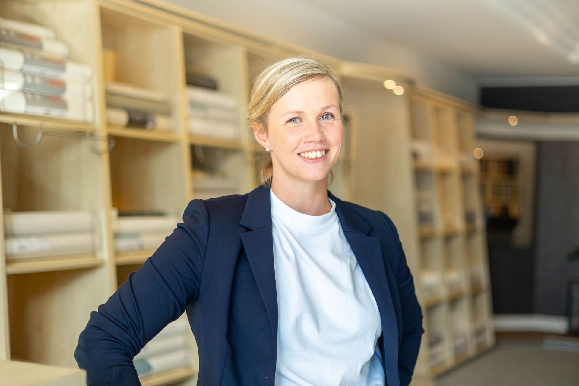 Eine Frau in blauer Jacke und weißem Hemd steht vor einer Regalwand.