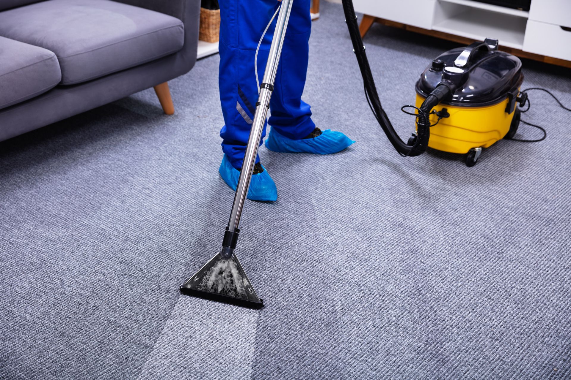Una persona con un mono azul limpia una alfombra gris con una aspiradora amarilla.