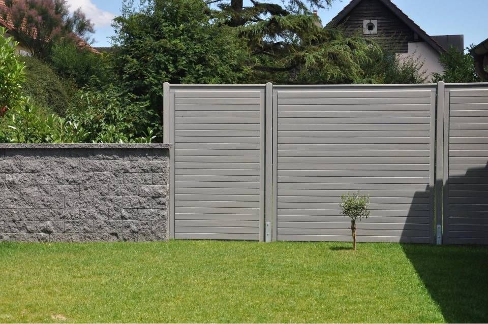 Clôture en lattes grises et muret en pierre dans une cour herbeuse, avec un petit arbre.
