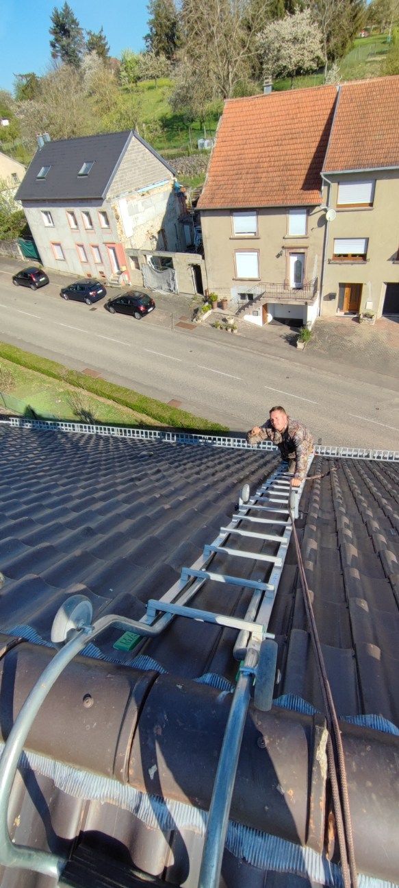 Une personne grimpe à une échelle posée sur un toit pentu de tuiles sombres, surplombant une rue bordée de bâtiments en pierre par une journée ensoleillée.
