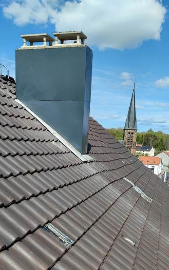 Vue depuis un toit de tuiles montrant une cheminée gris foncé avec deux chapeaux de conduit, se détachant sur le ciel et une église au loin.