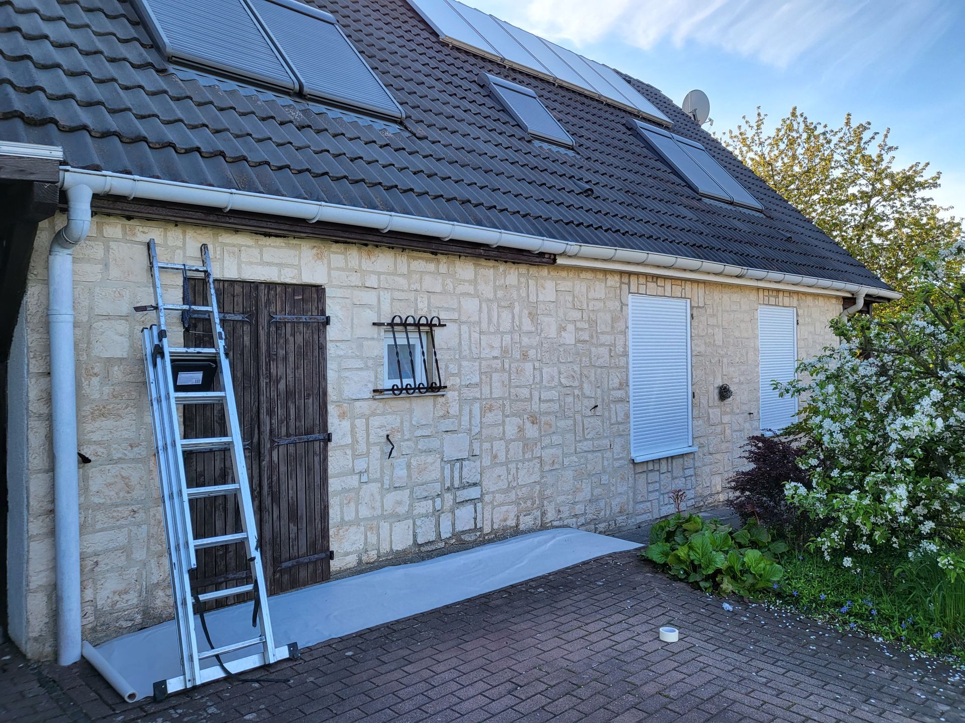 Une maison en pierre beige avec un toit sombre et des panneaux solaires, avec une échelle appuyée contre une porte en bois et deux fenêtres à volets.