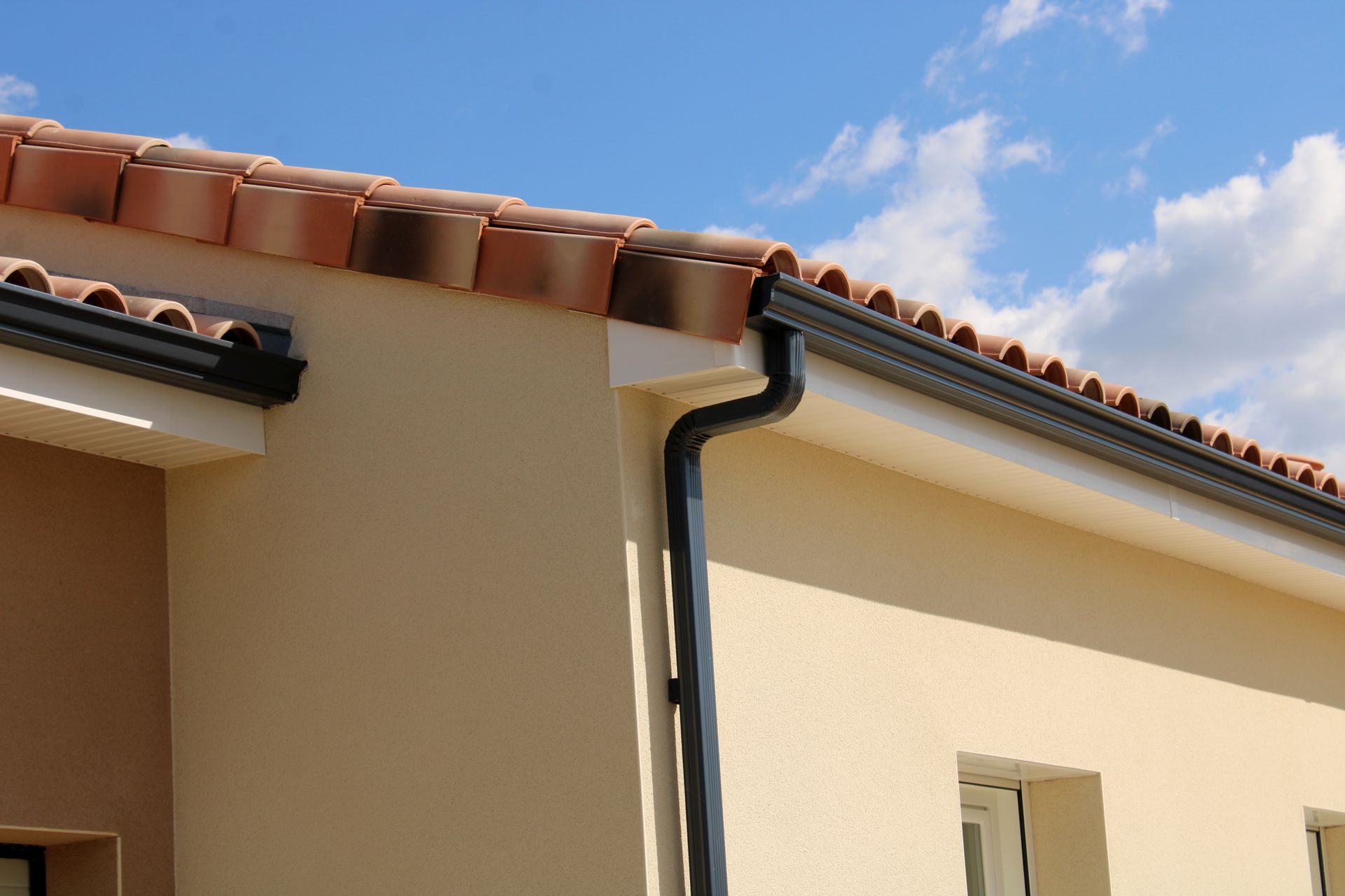 Un bâtiment à l'extérieur beige, avec un toit de tuiles en terre cuite, des gouttières noires et une descente d'eau pluviale, se détachant sur un ciel bleu nuageux.