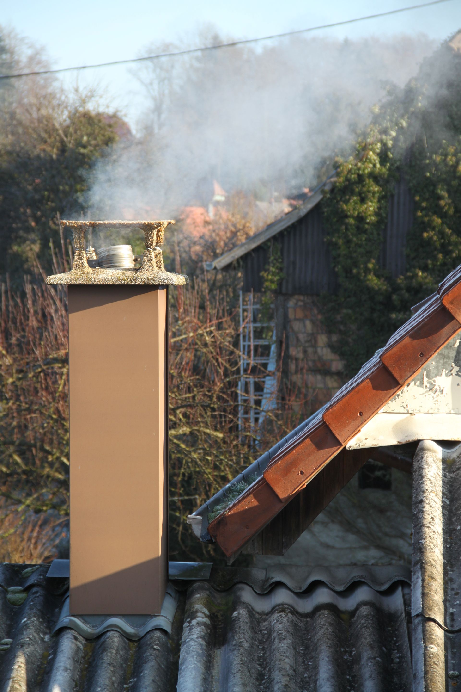 Une cheminée beige fumante sur un toit en tôle ondulée, à côté d'un toit de tuiles, avec des arbres en arrière-plan.