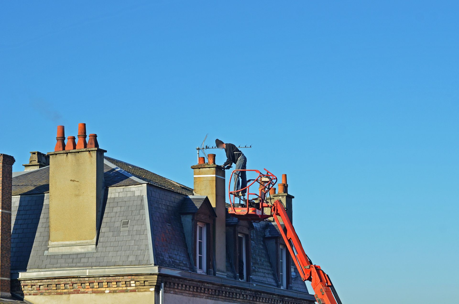 Un ouvrier, à bord d'une nacelle élévatrice rouge, effectue des travaux d'entretien sur une cheminée au sommet d'un toit en pente, sur fond de ciel bleu dégagé.