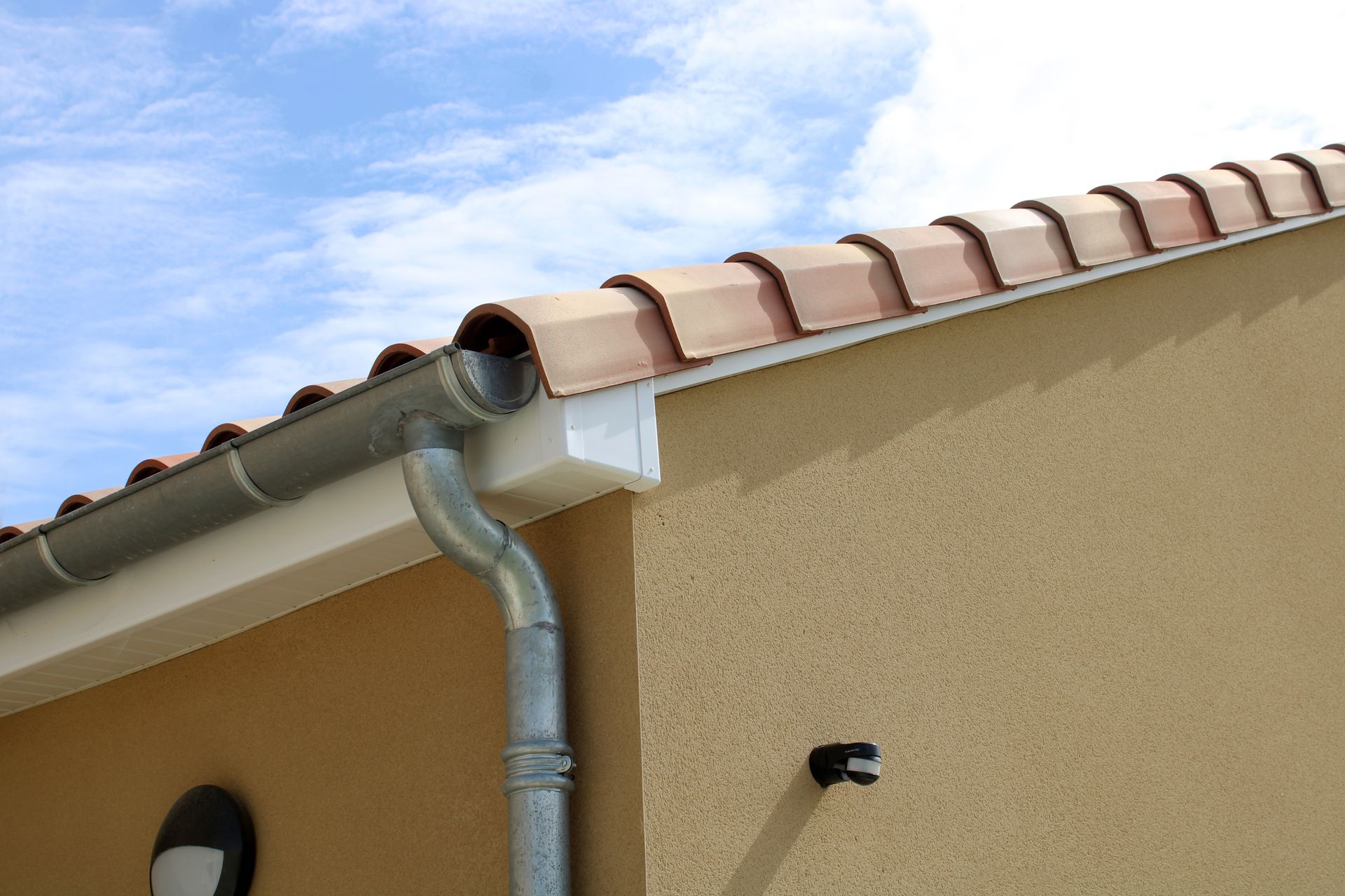 Une gouttière et un tuyau de descente en métal fixés au bord d'une maison beige au toit de tuiles en terre cuite, sous un ciel bleu.