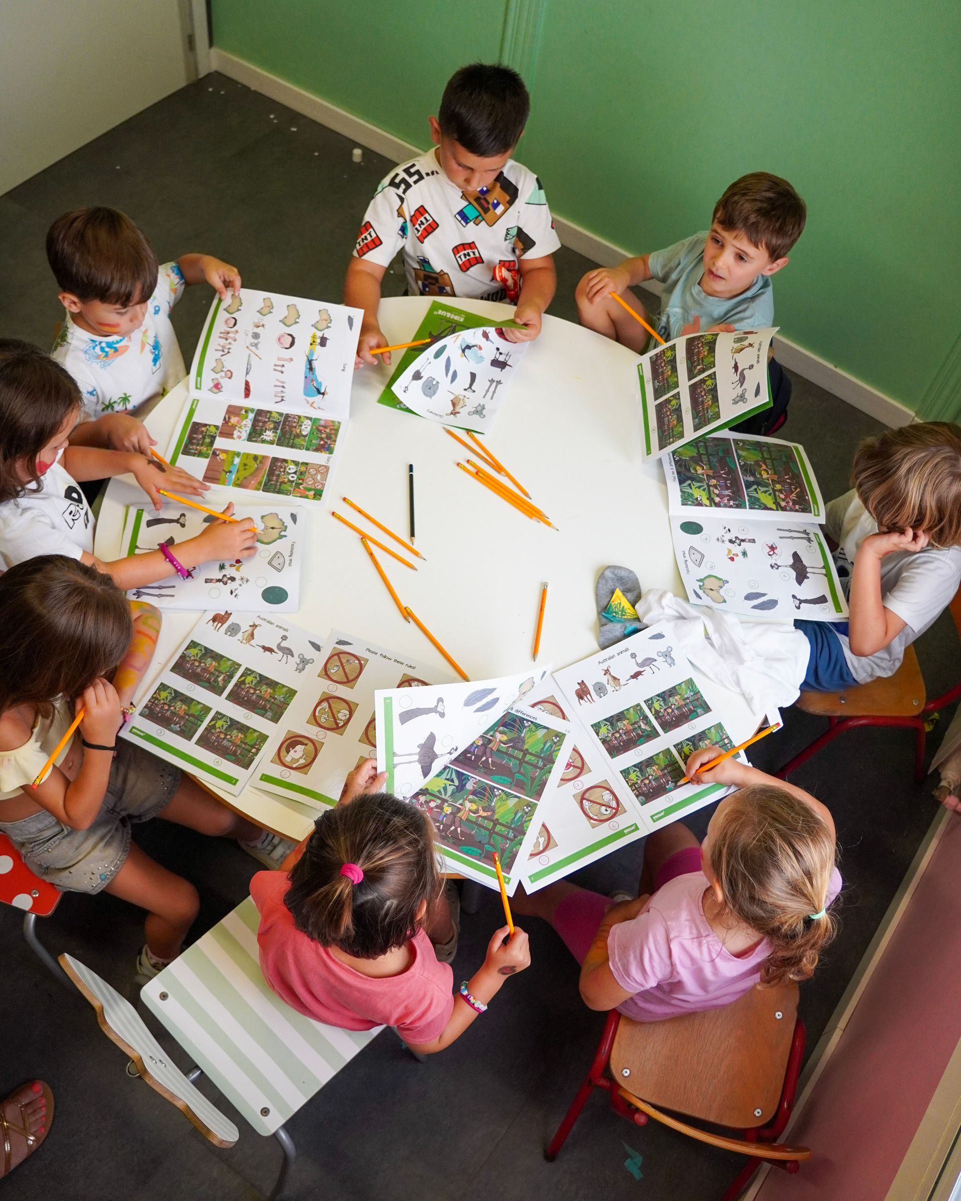 Niños sentados alrededor de una mesa redonda, leyendo libros ilustrados y usando lápices en un aula.