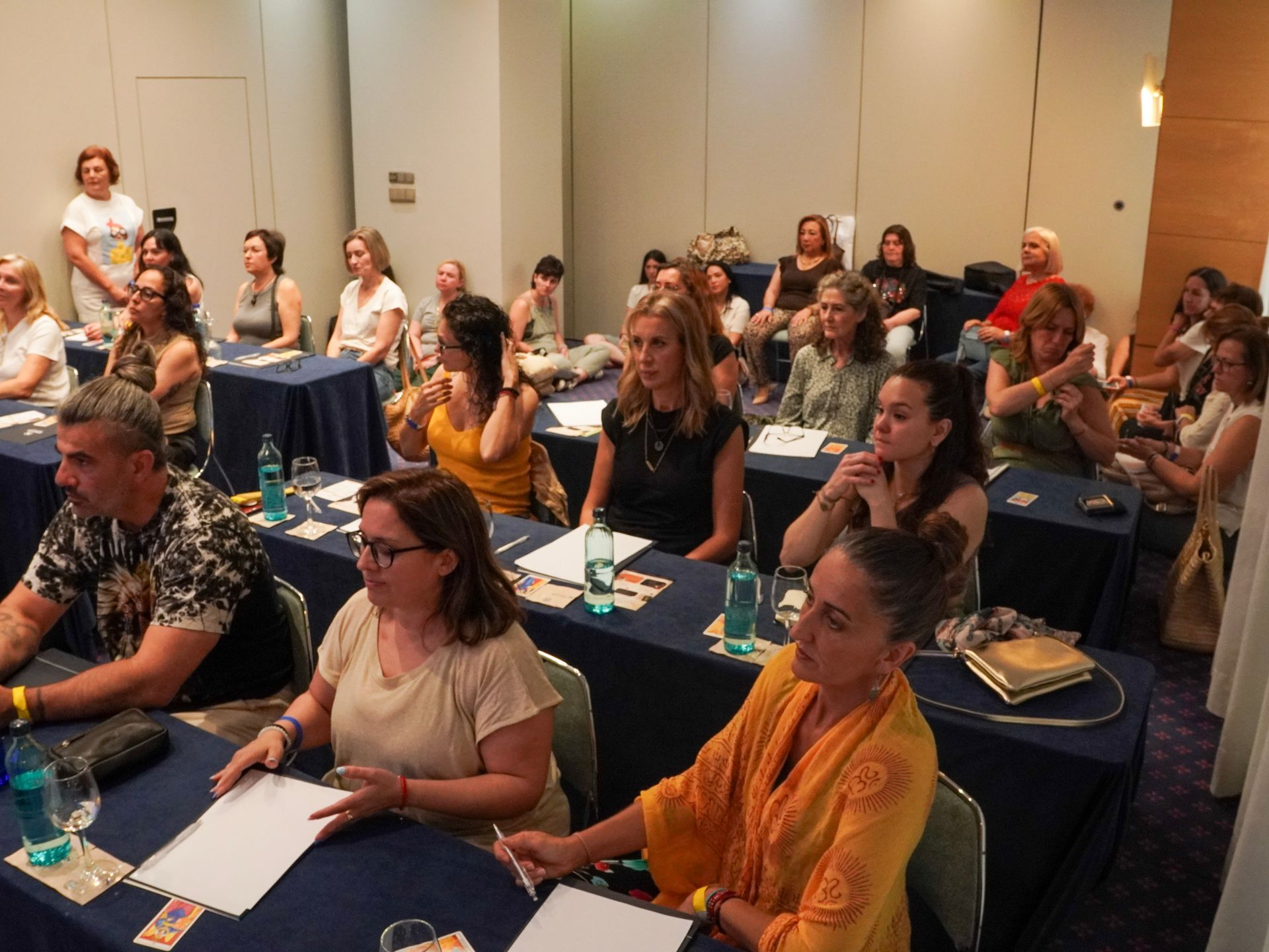 El público, sentado en una sala de conferencias, escucha atentamente en mesas con cuadernos y botellas de agua.