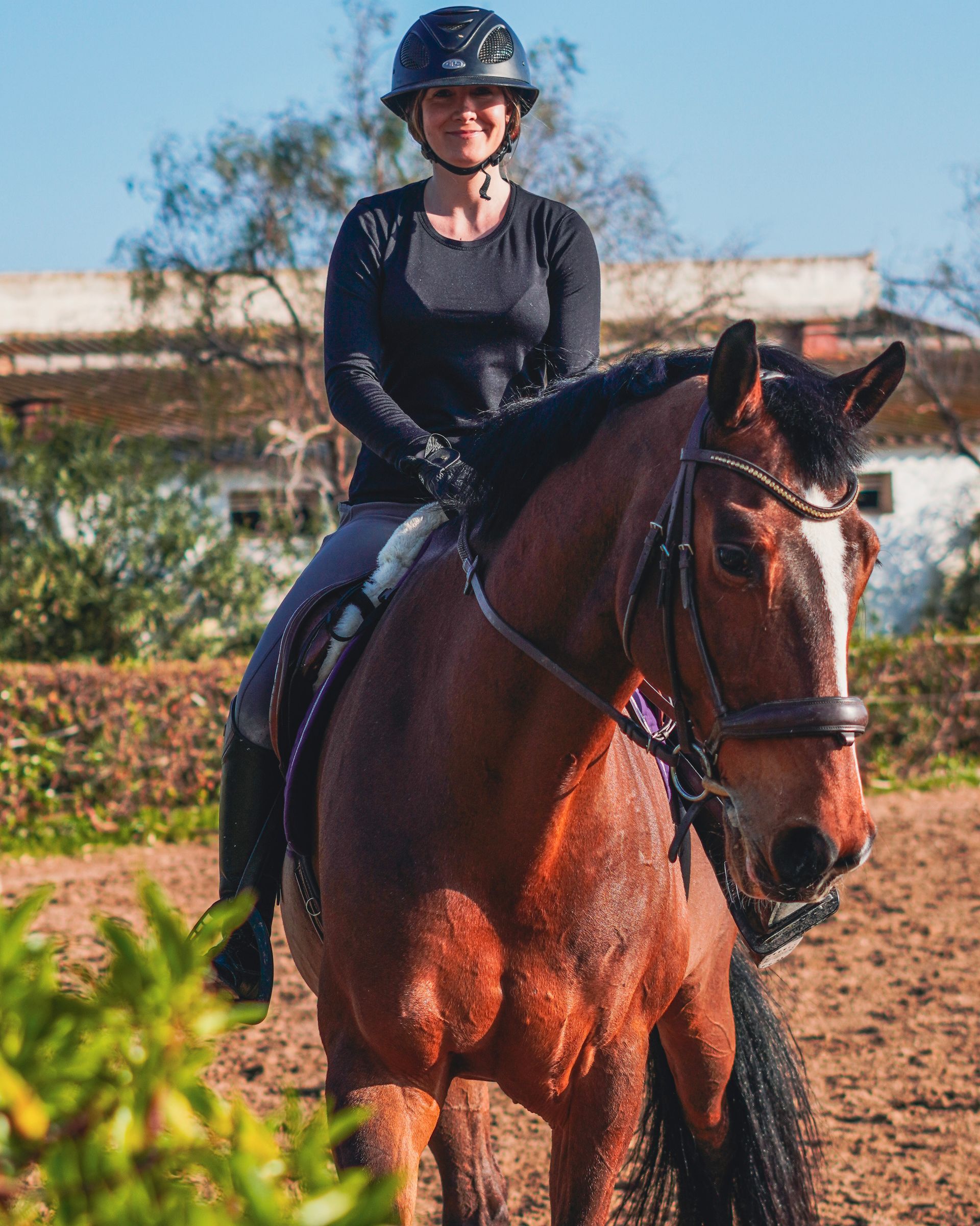 Persona con atuendo de equitación negro sobre un caballo marrón al aire libre en un día soleado.