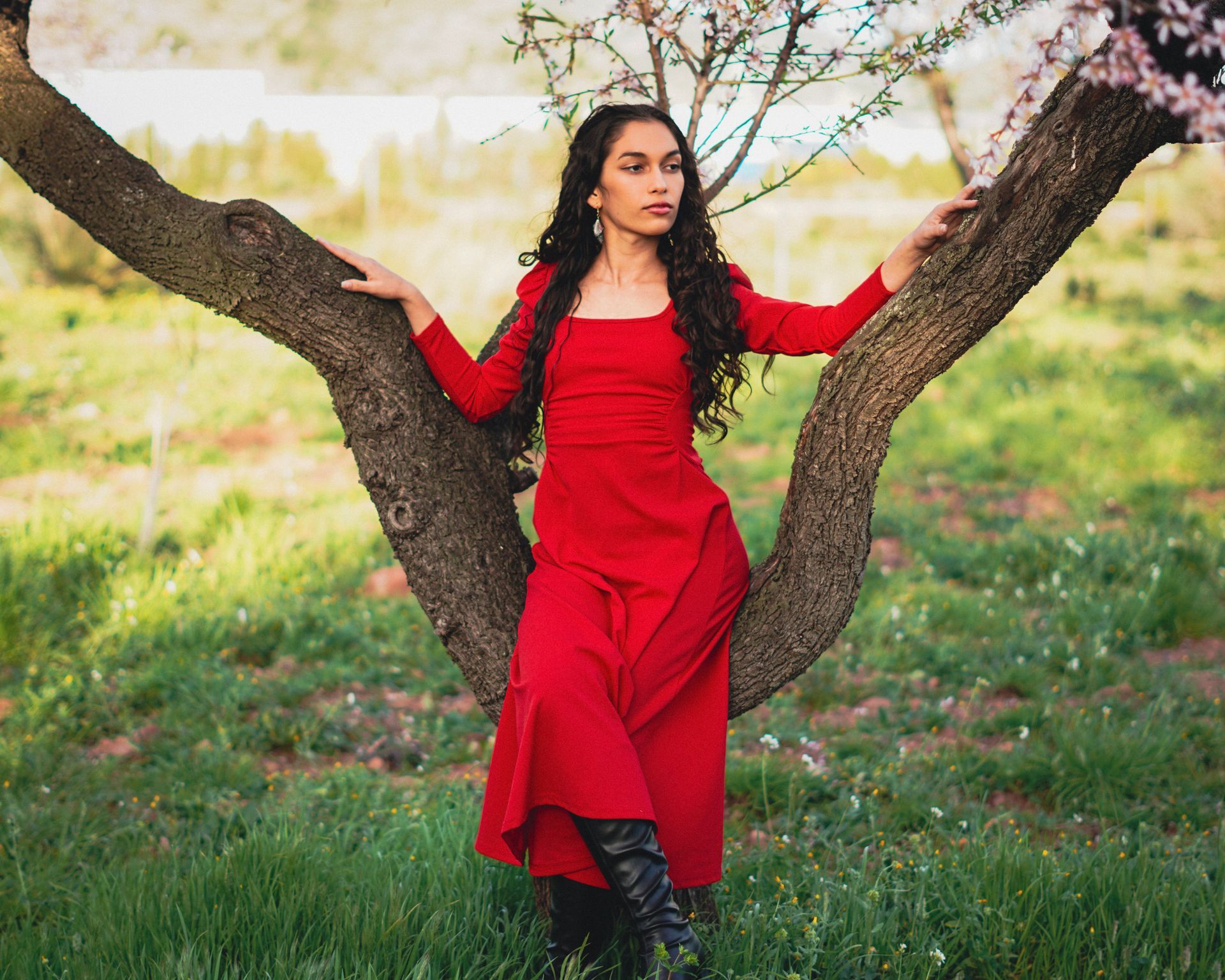 Mujer con un vestido rojo de pie entre dos troncos de árboles en un entorno exterior cubierto de hierba.
